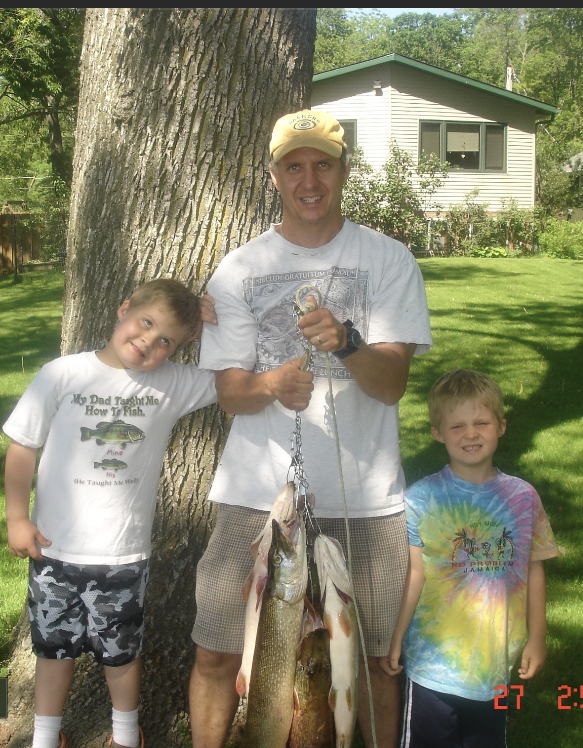 A man and two young boys standing outdoors near a tree, holding fishing catch with the man in the middle holding a chain with fish, in a backyard with a house in the background.