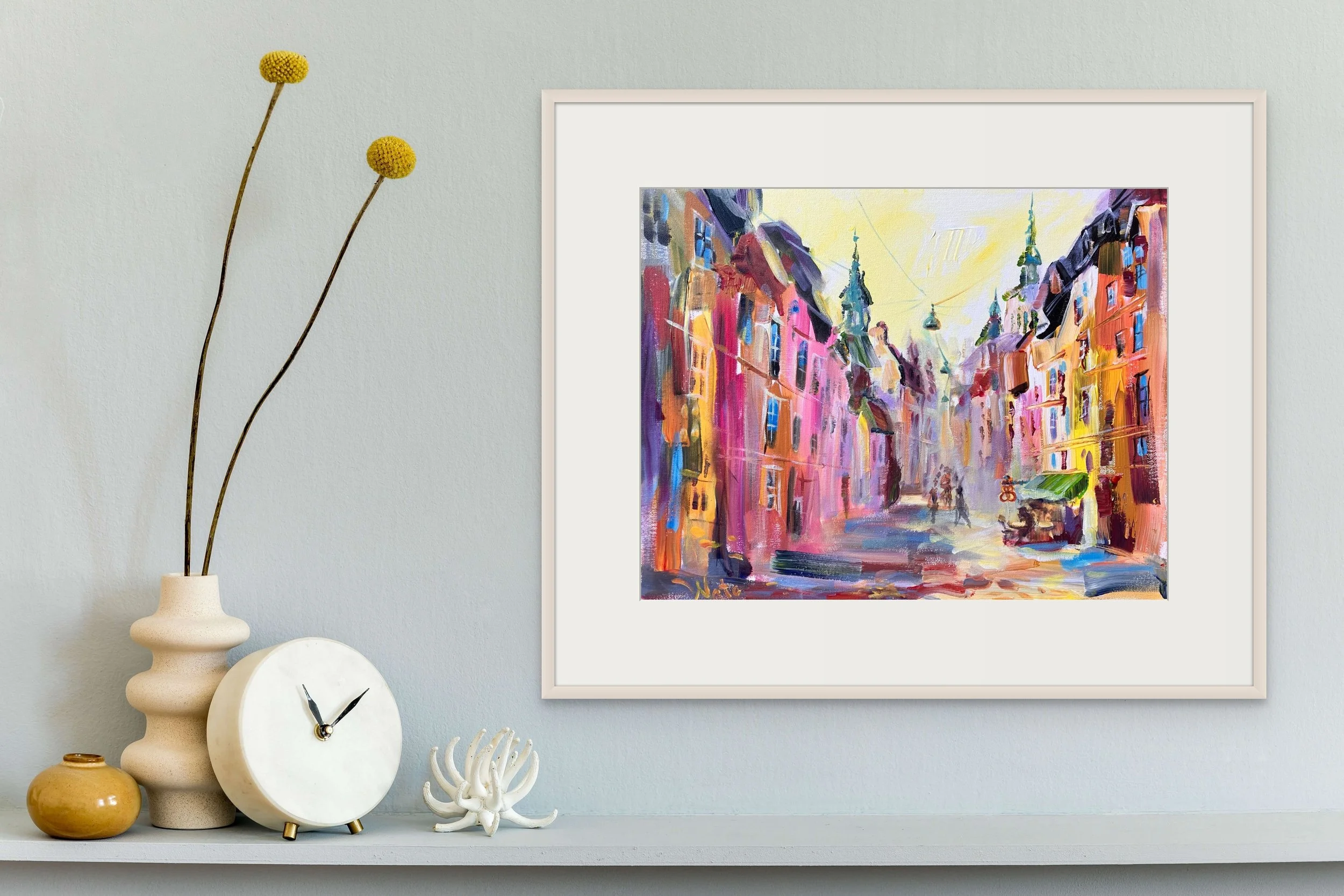 A colorful street scene painting hung on a light green wall, with a white shelf below holding a white clock, a beige vase with two long yellow flowers, a textured orange vase, and a white decorative coral piece.