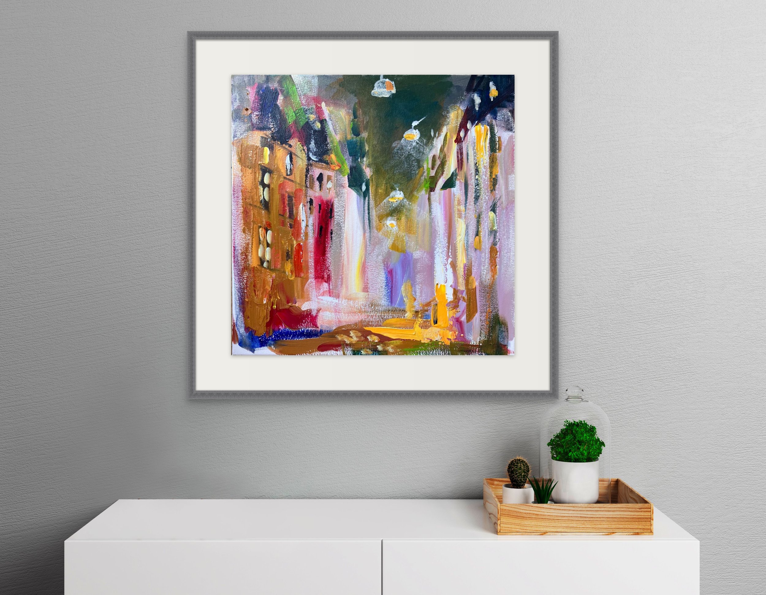 Abstract cityscape painting with colorful buildings and street scene, hung on a gray wall above a white sideboard with potted plants and a wooden tray.