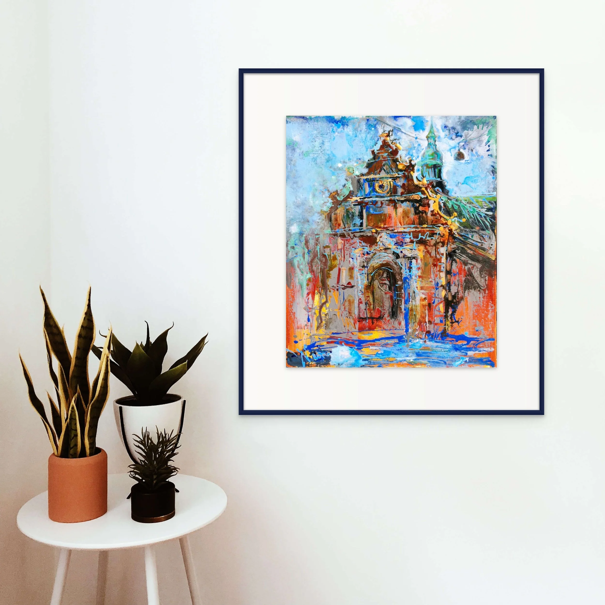Colorful abstract painting of a historical building, featuring a prominent archway and domed towers, mounted on a white wall, with a small white table below holding three potted plants.