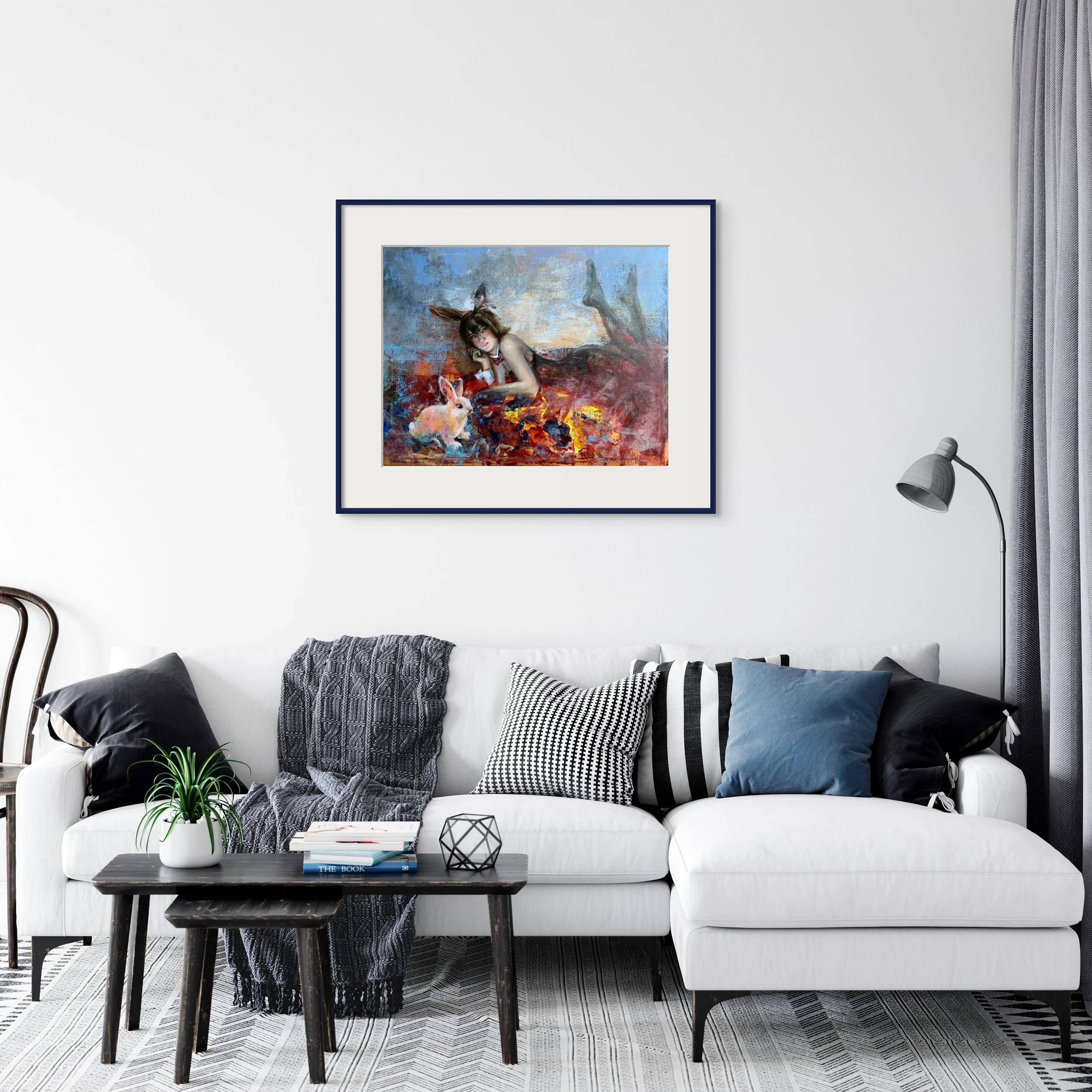 Living room with a white sofa, decorative pillows, a dark wood coffee table with books and a small green plant, a black side table, a floor lamp, gray curtains, and a framed colorful painting of a girl with a rabbit.