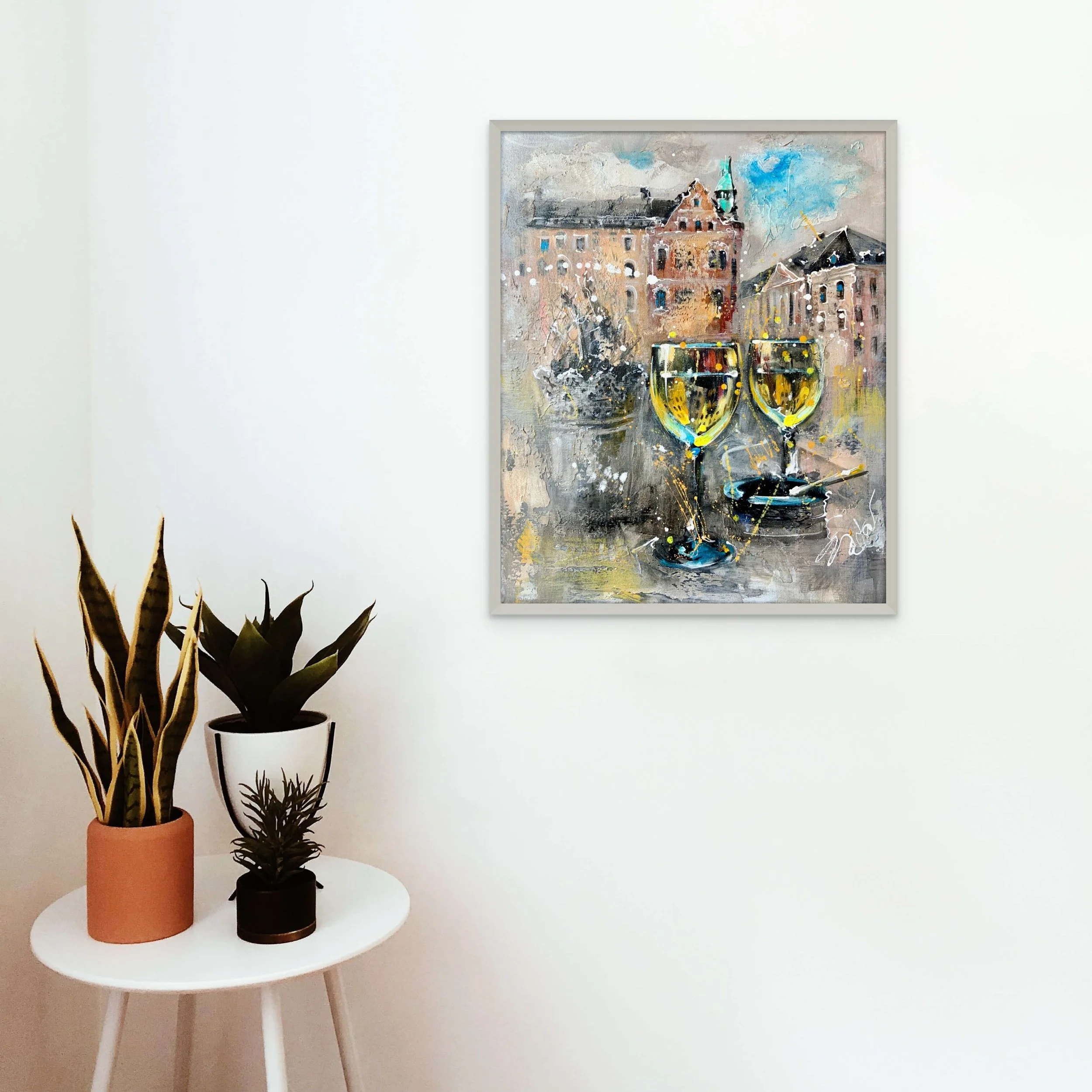 A modern interior with a white wall, a framed abstract painting of city buildings and two glasses of wine, and a small round white table holding three potted plants.