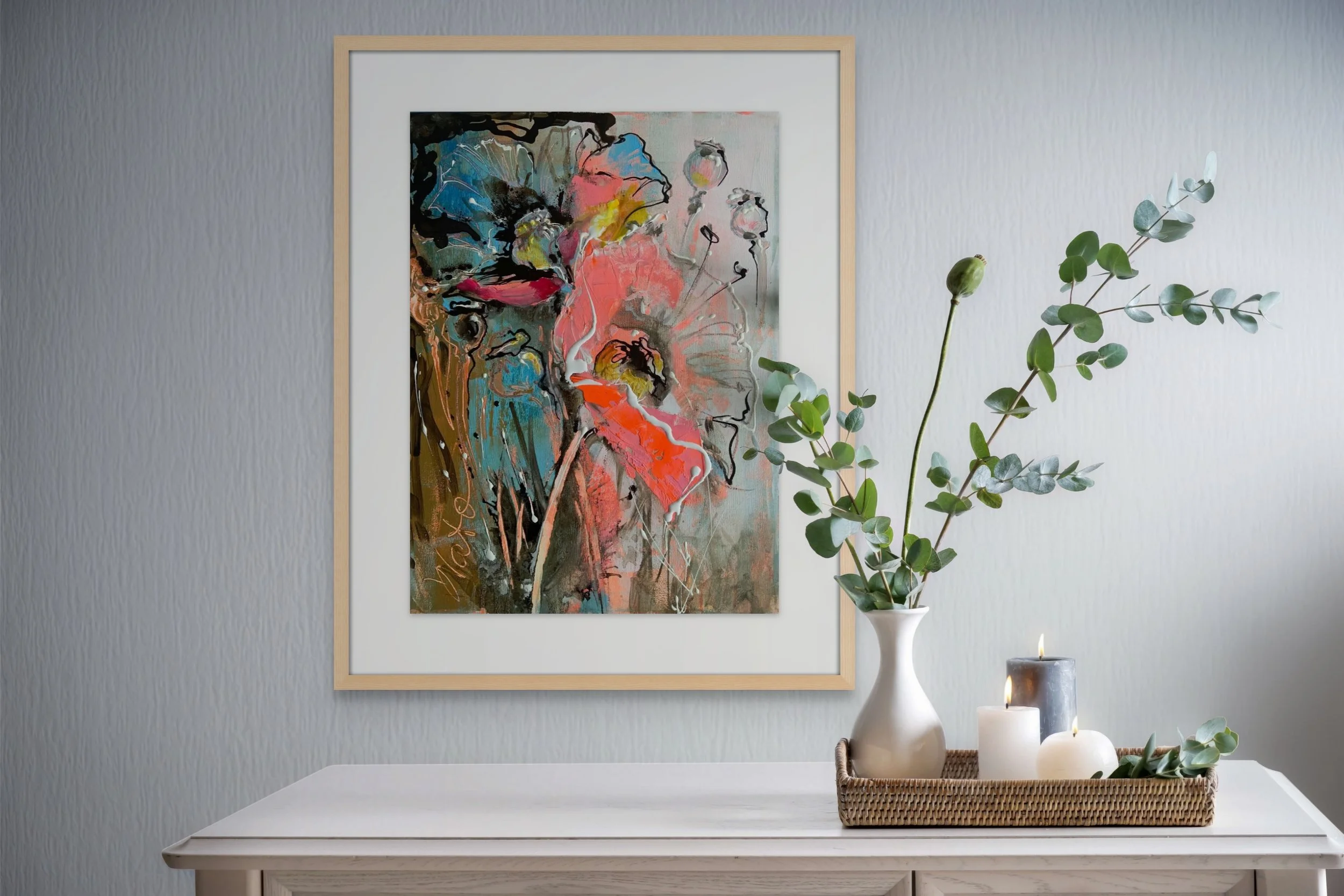 Decorative interior scene featuring an abstract floral painting on the wall, a white vase with green eucalyptus branches, and a tray with candles on a white wooden surface.