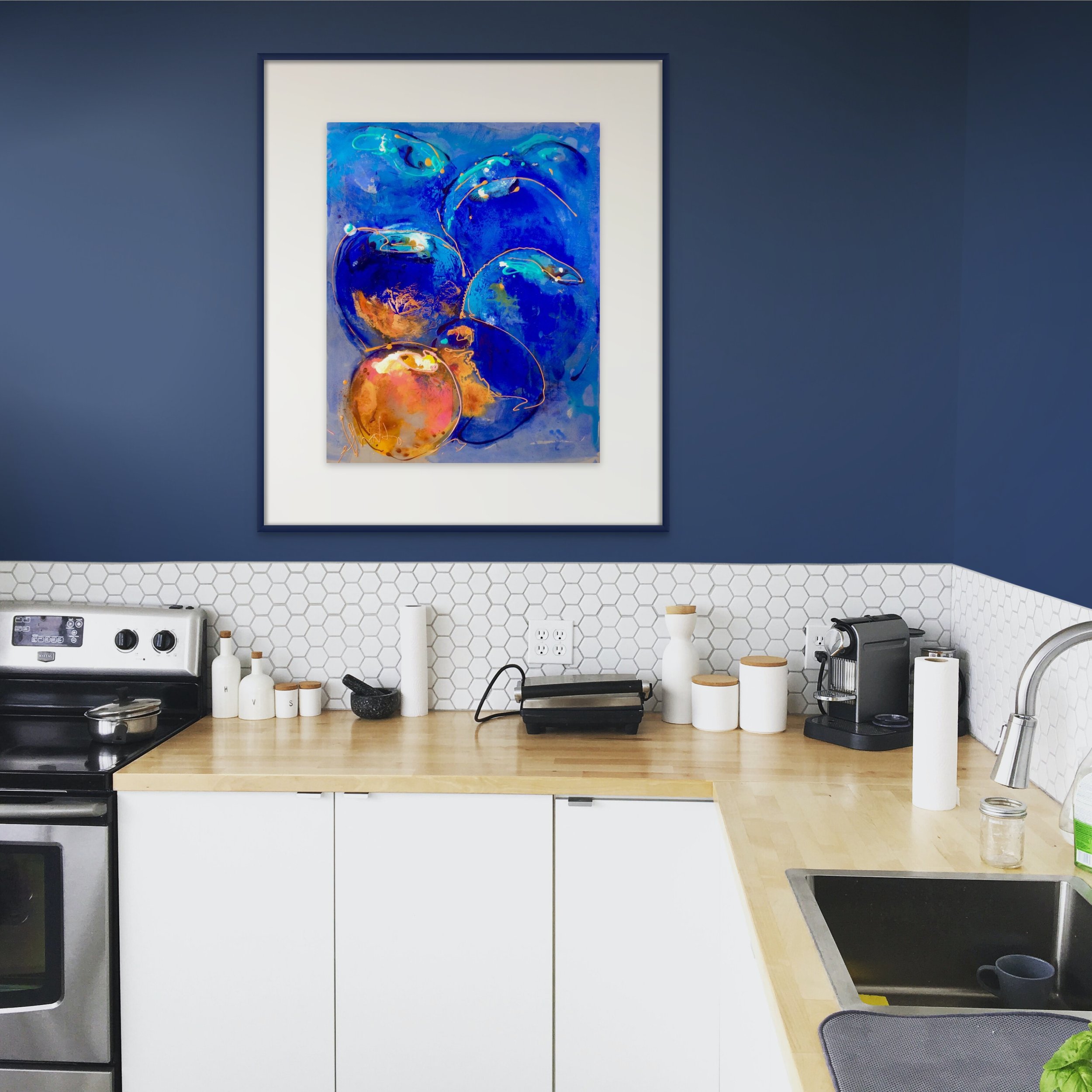 A kitchen with a blue wall and white hexagon tile backsplash. A colorful abstract painting of blue and orange circles hangs above the countertop.