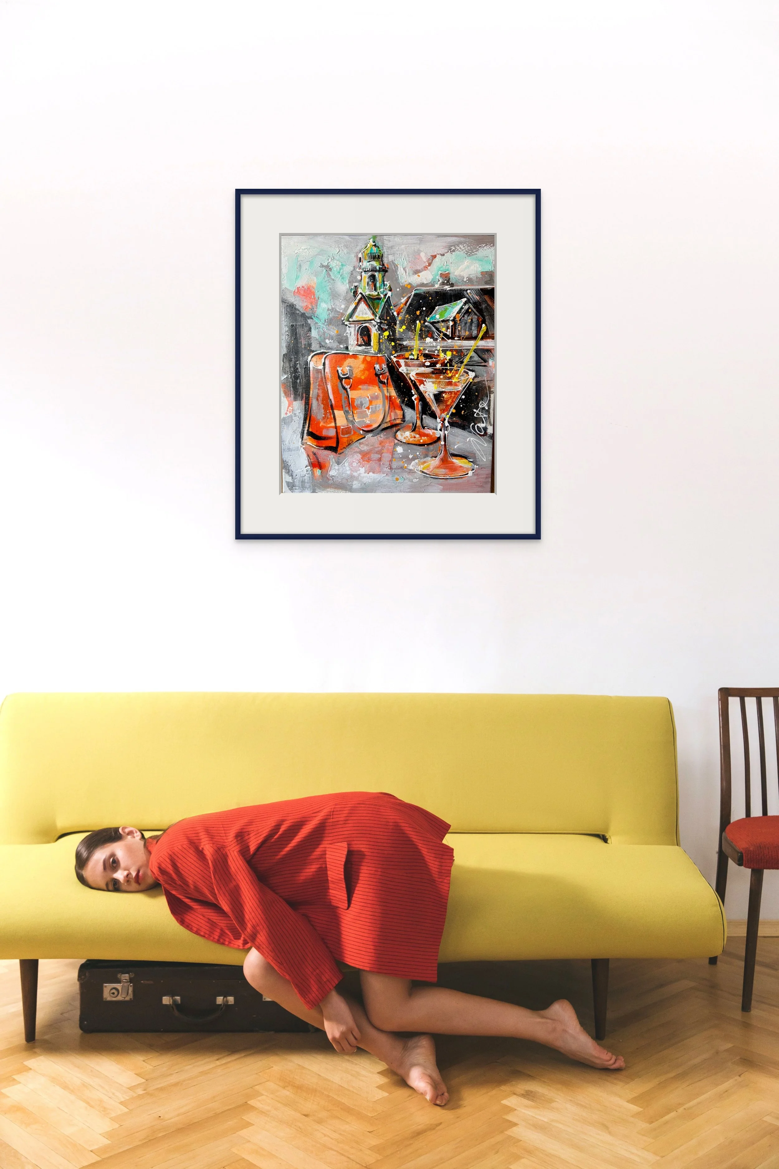 A woman in a red dress is lying on her side on a yellow sofa, with her head resting on the armrest and feet on the floor. There is a black suitcase under the sofa. A framed colorful abstract painting hangs on the wall above, featuring a church, table