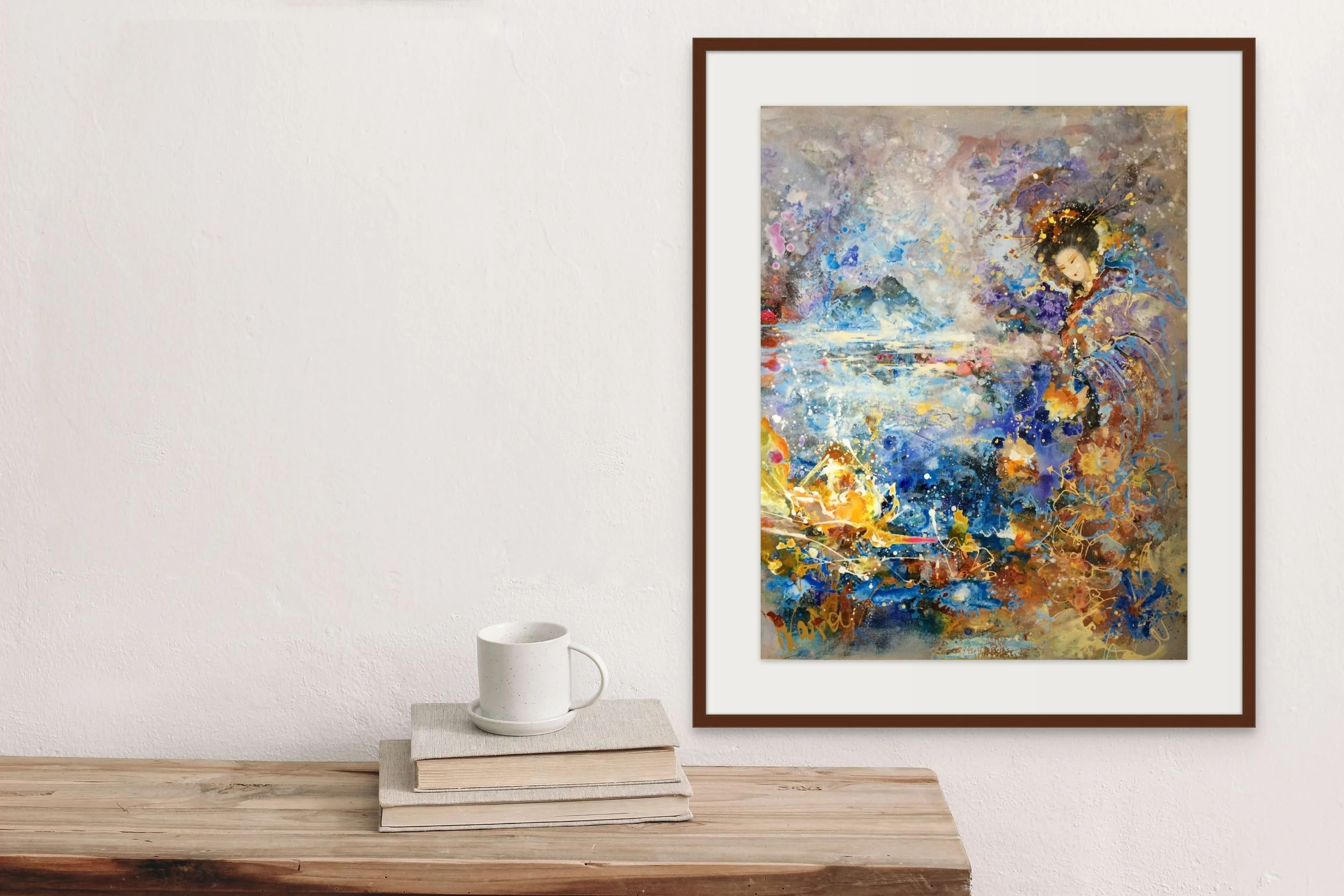 Colorful abstract watercolor painting of a woman in traditional dress hanging on a white wall with a wooden frame, placed beside a wooden table with a stack of books and a white mug.