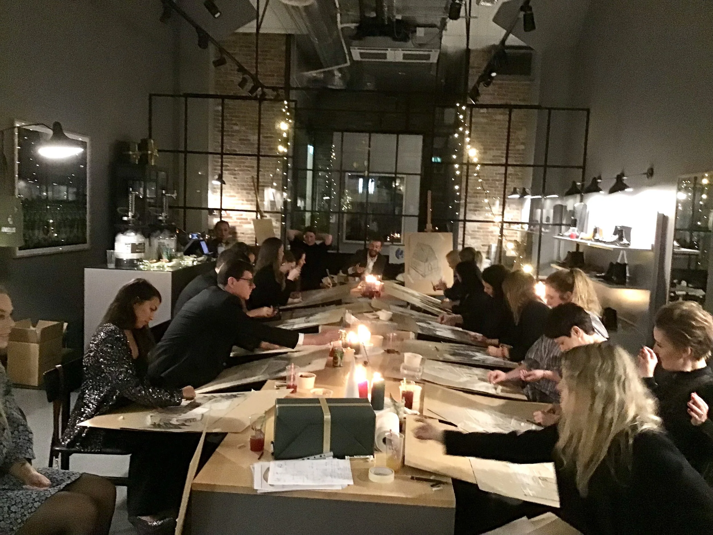 Kursister hos Nata Watts sidder ved et langt bord i et hyggeligt, indrettet rum med bløde lys fra stearinlys og julelys, og arbejder med papir og skitser.