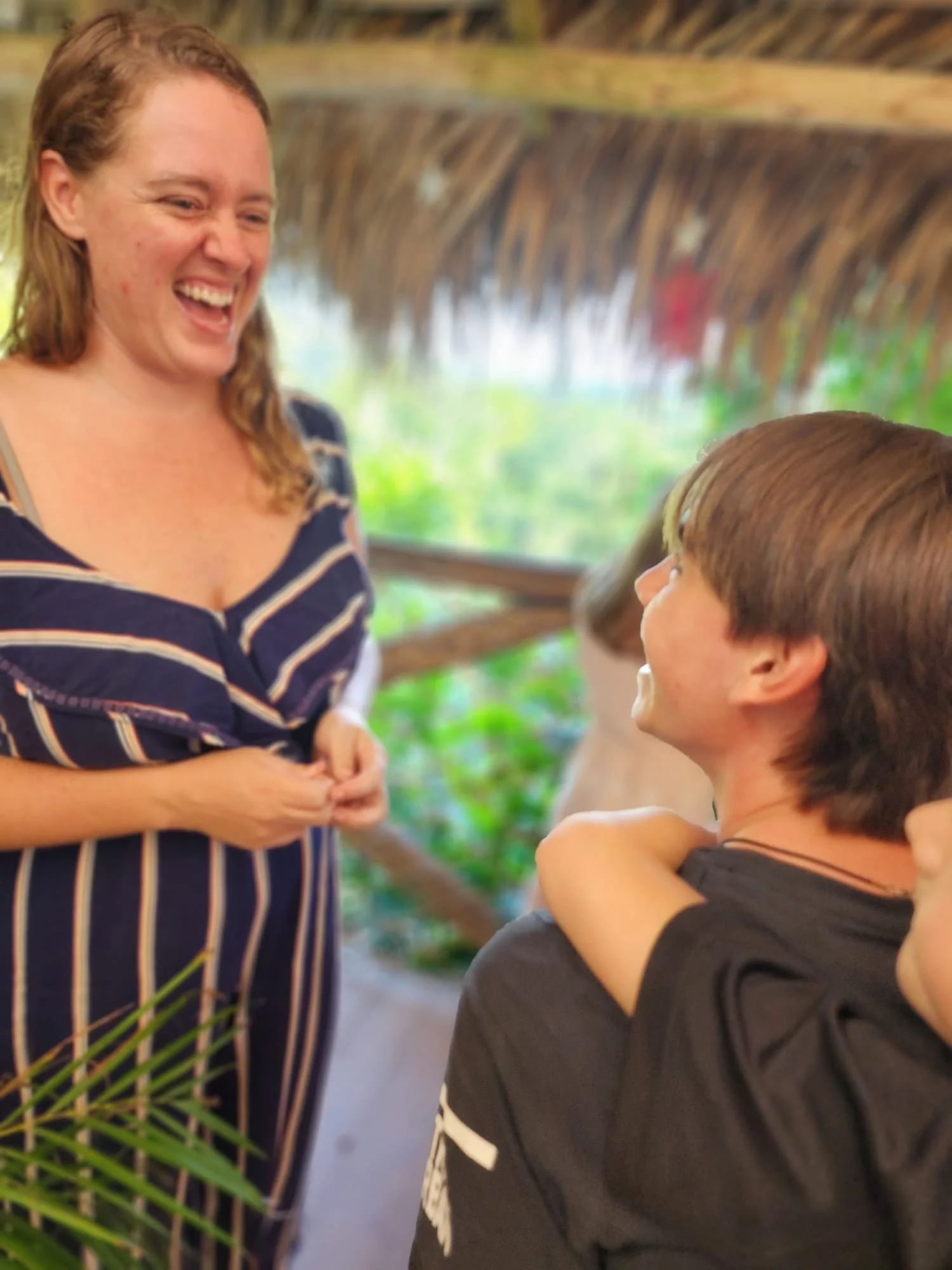 A woman and a boy are smiling and laughing at each other in an outdoor setting with lush greenery and a thatched roof overhead.