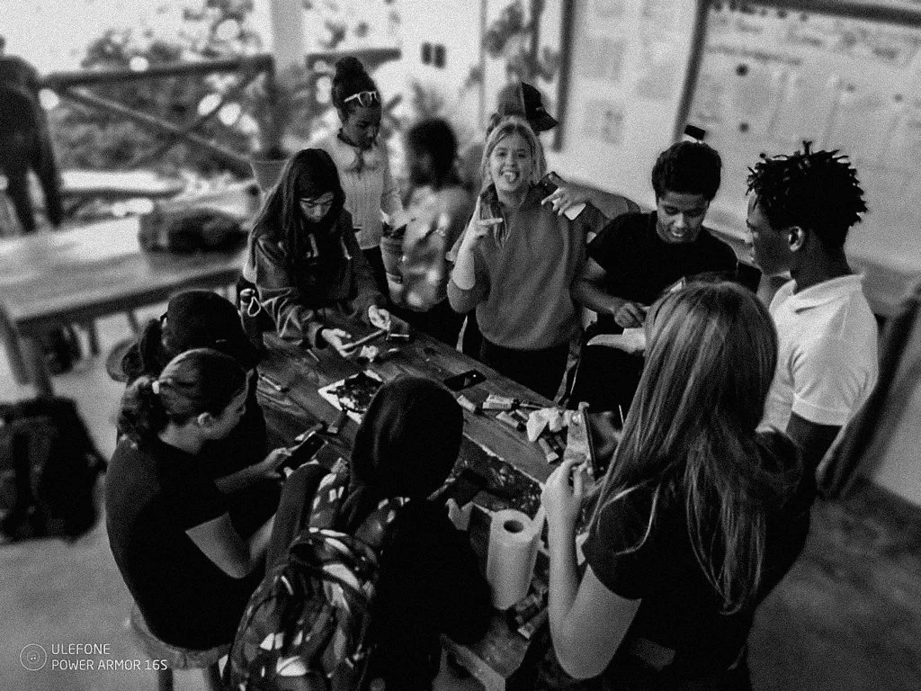 Group of children gathered around a table, engaging in an activity, with some using electronic devices, in a classroom or workshop setting.