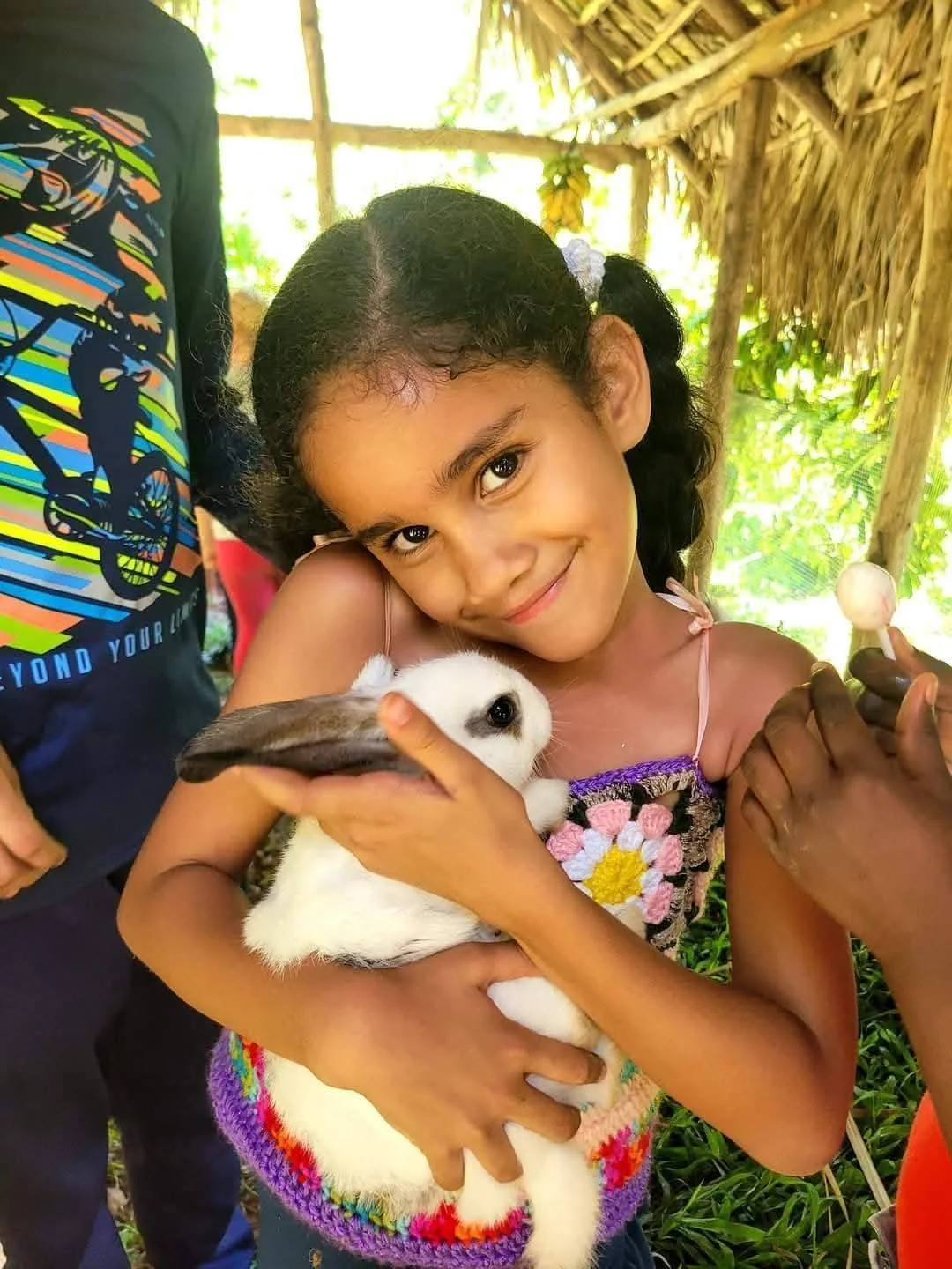 A young girl with dark hair and a colorful, flower-embroidered dress, smiling while holding a white rabbit with black markings. The girl is in a wooden shelter with green plants in the background, and a hand of an adult is visible near her holding a 
