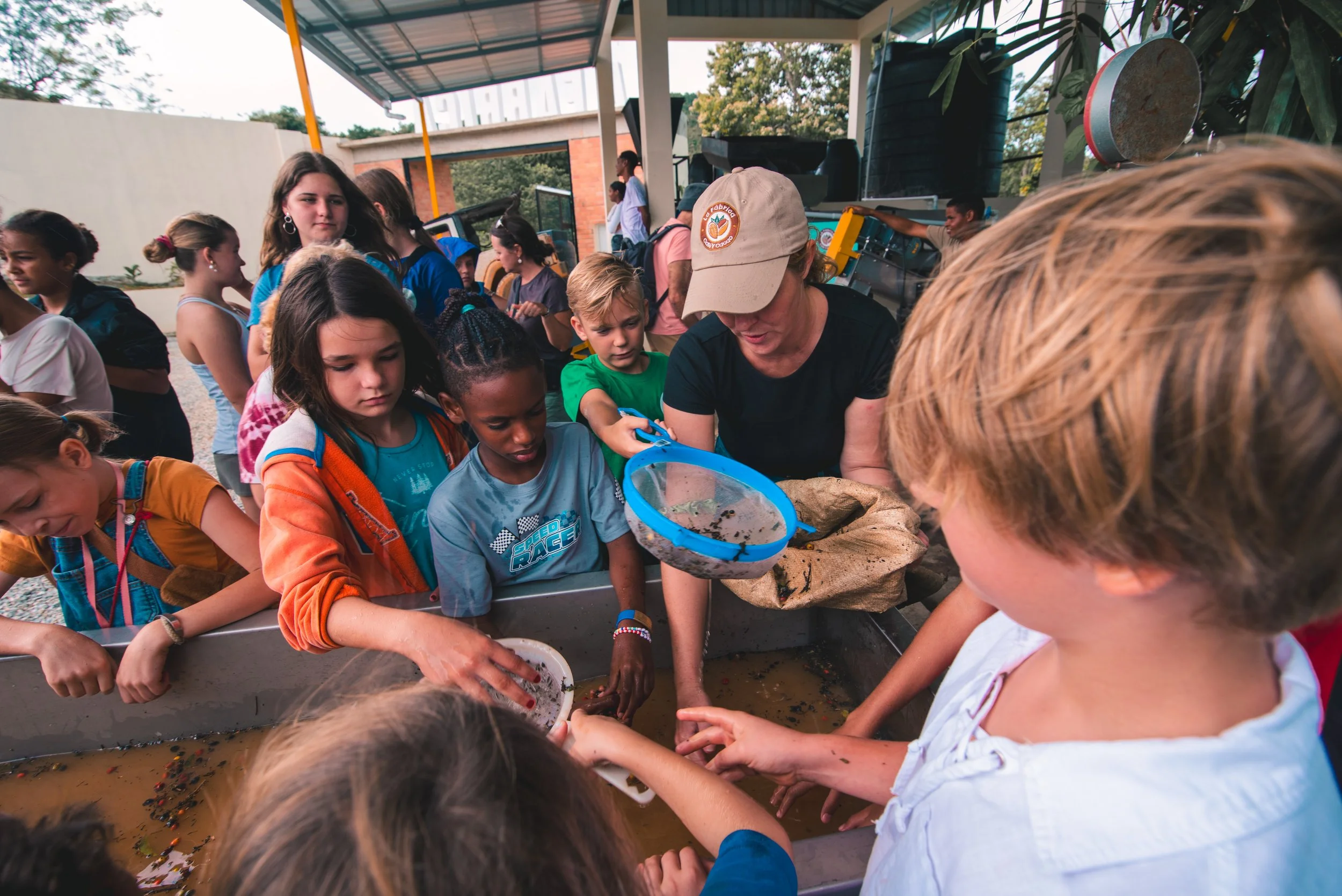 Group of children and adults at a scientific exhibit, examining a rectangular tank with water and small objects or creatures, outdoors under a shelter.