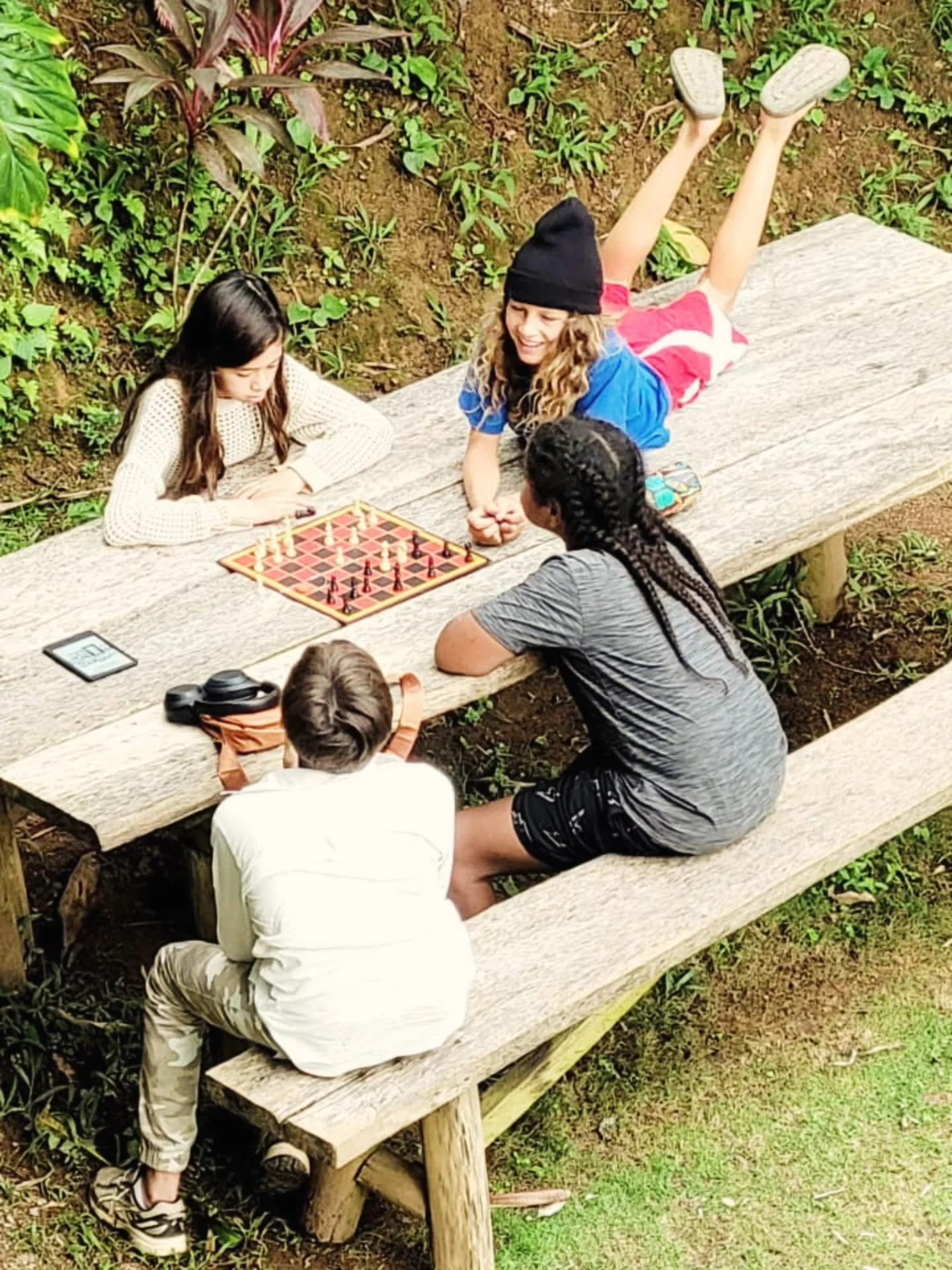 Five children are playing chess on a wooden outdoor table surrounded by greenery.