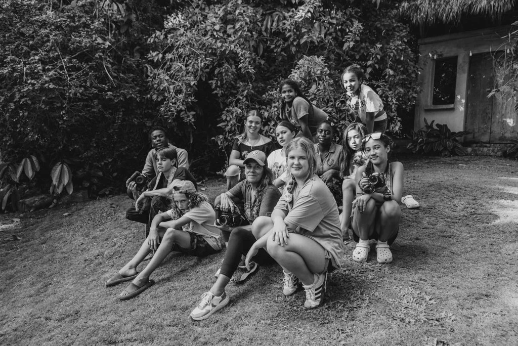 Group of children and teens outdoors, some sitting and others standing, in a shaded area with trees and bushes, posing for a photograph.