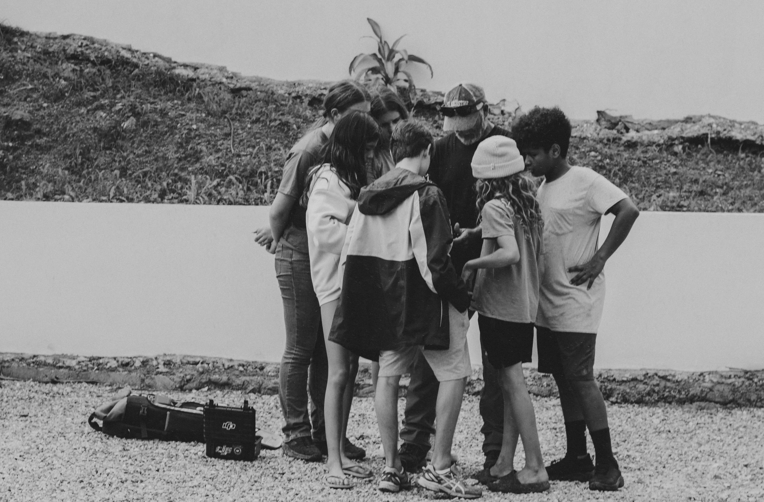 A group of people standing together outdoors, looking at something on a phone, with a backpack on the ground nearby.