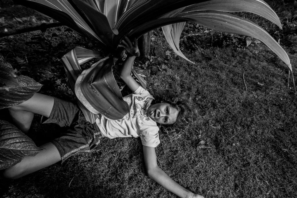 A young girl with curly hair lying on the ground next to large plant leaves, looking surprised or shocked.