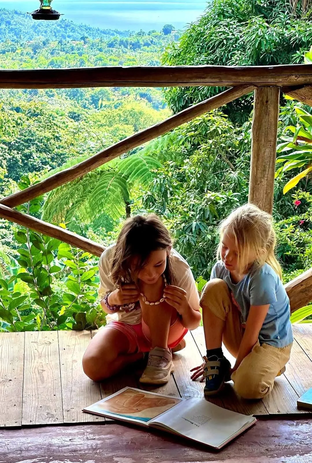 Two children sitting on wooden floor reading a book, surrounded by lush green tropical foliage, with hills in the background.