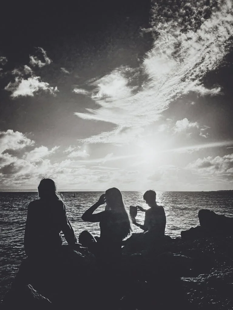 Silhouettes of three people sitting on rocks by the water at sunset with a cloudy sky.