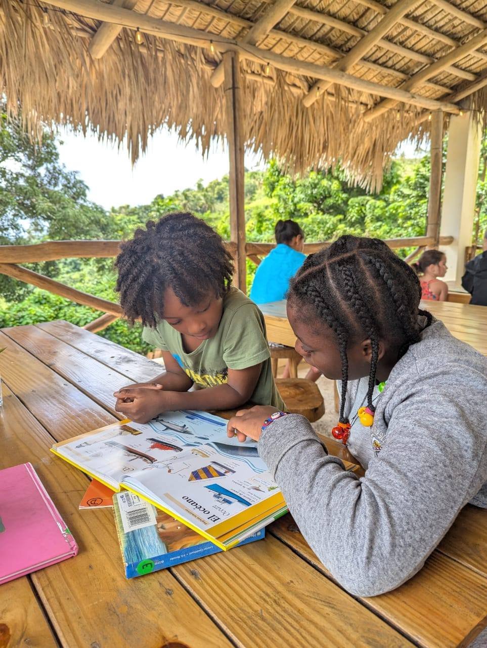 Two children looking at an open book on a wooden table under a thatched roof, with other children in the background sitting on wooden benches, surrounded by green trees.