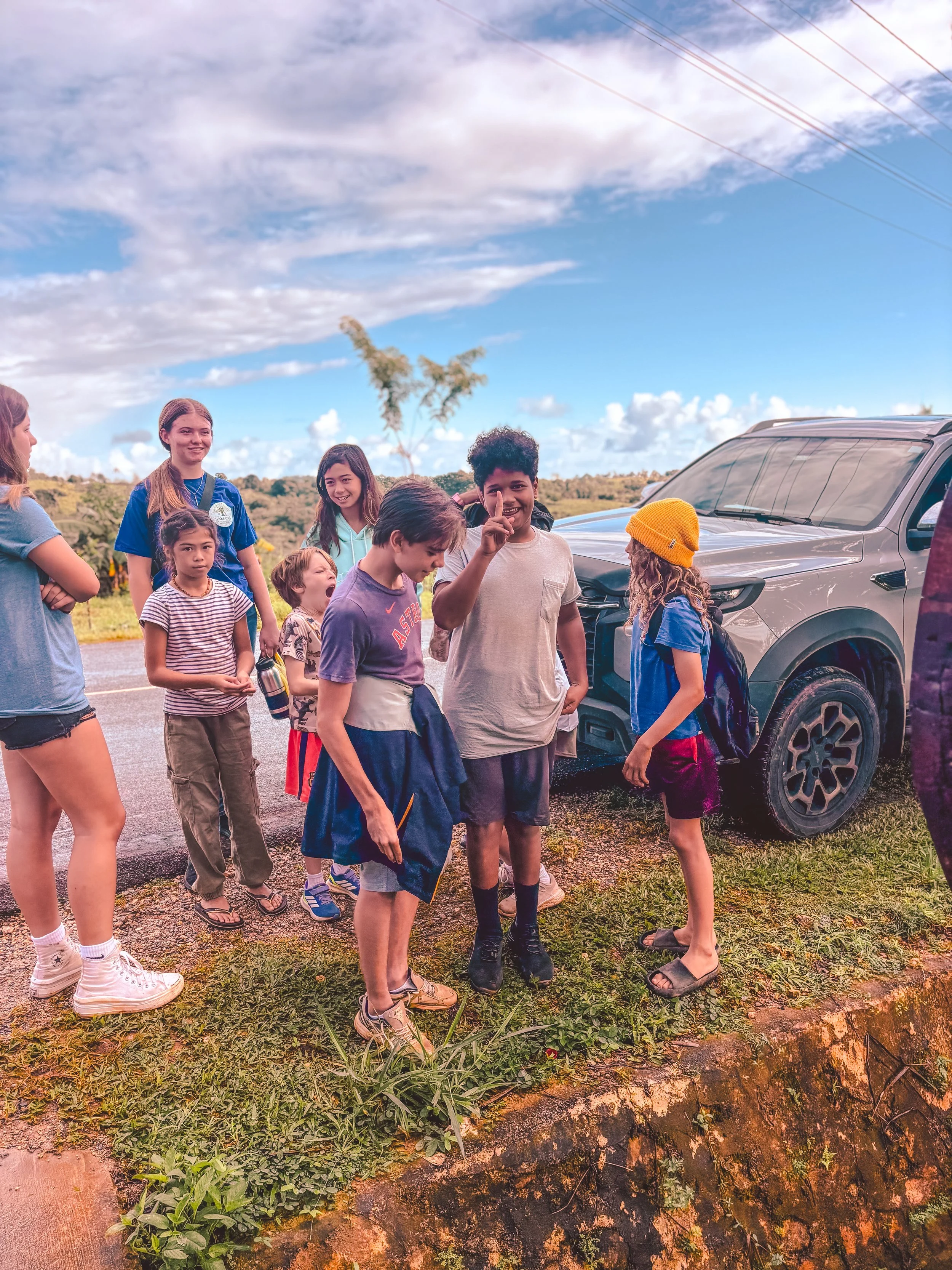 Group of children and young adults standing near a silver SUV outdoors on a cloudy day with power lines in the background.