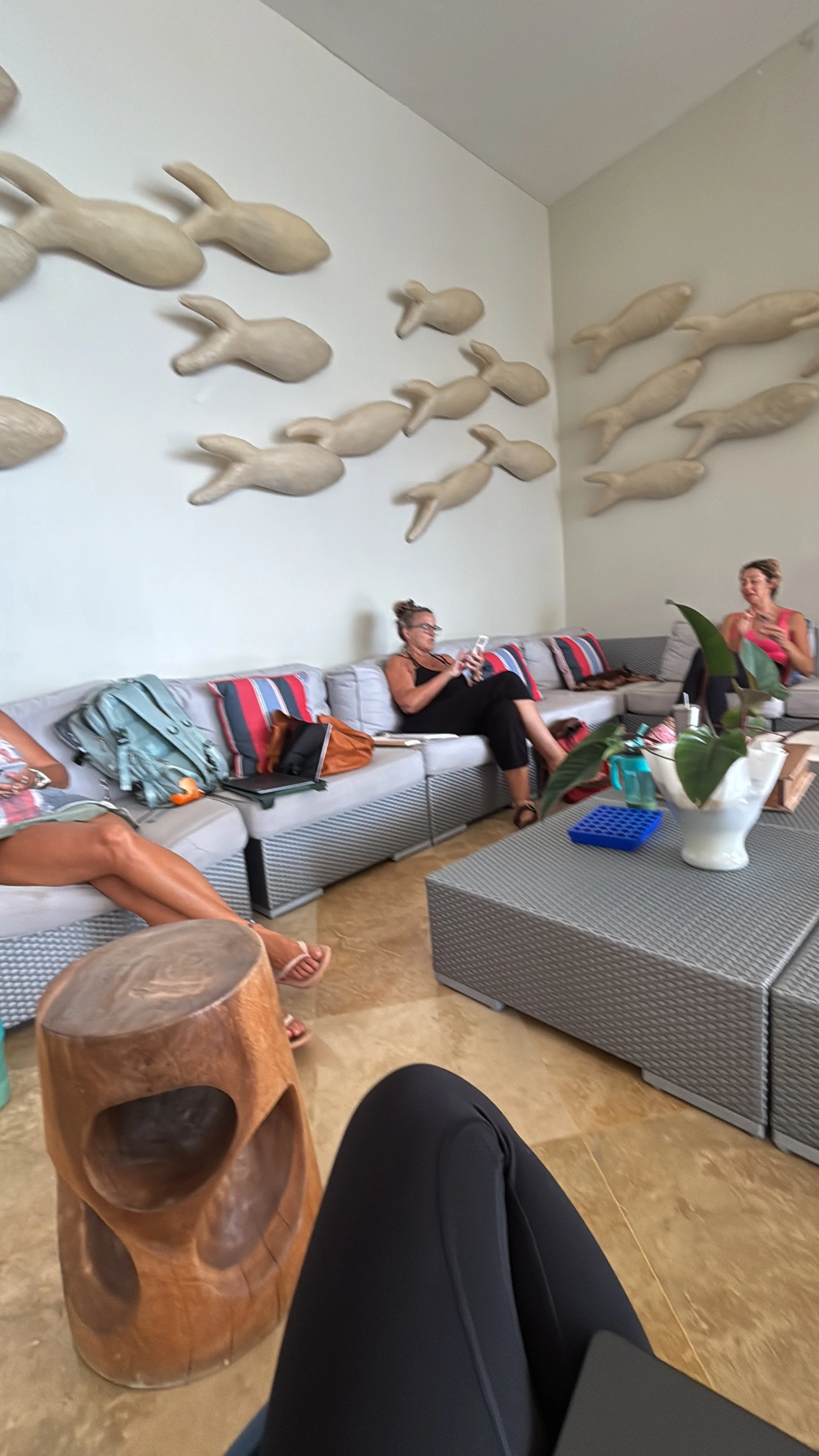 People sitting on a gray couch in a room with whale sculptures on the wall, some with bags and laptops, a wooden stool in the foreground, and a table with plants and various items.