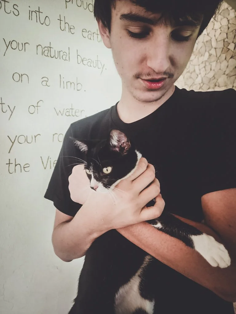 A young man holding a black and white cat indoors with a decorative wall and handwritten text in the background.