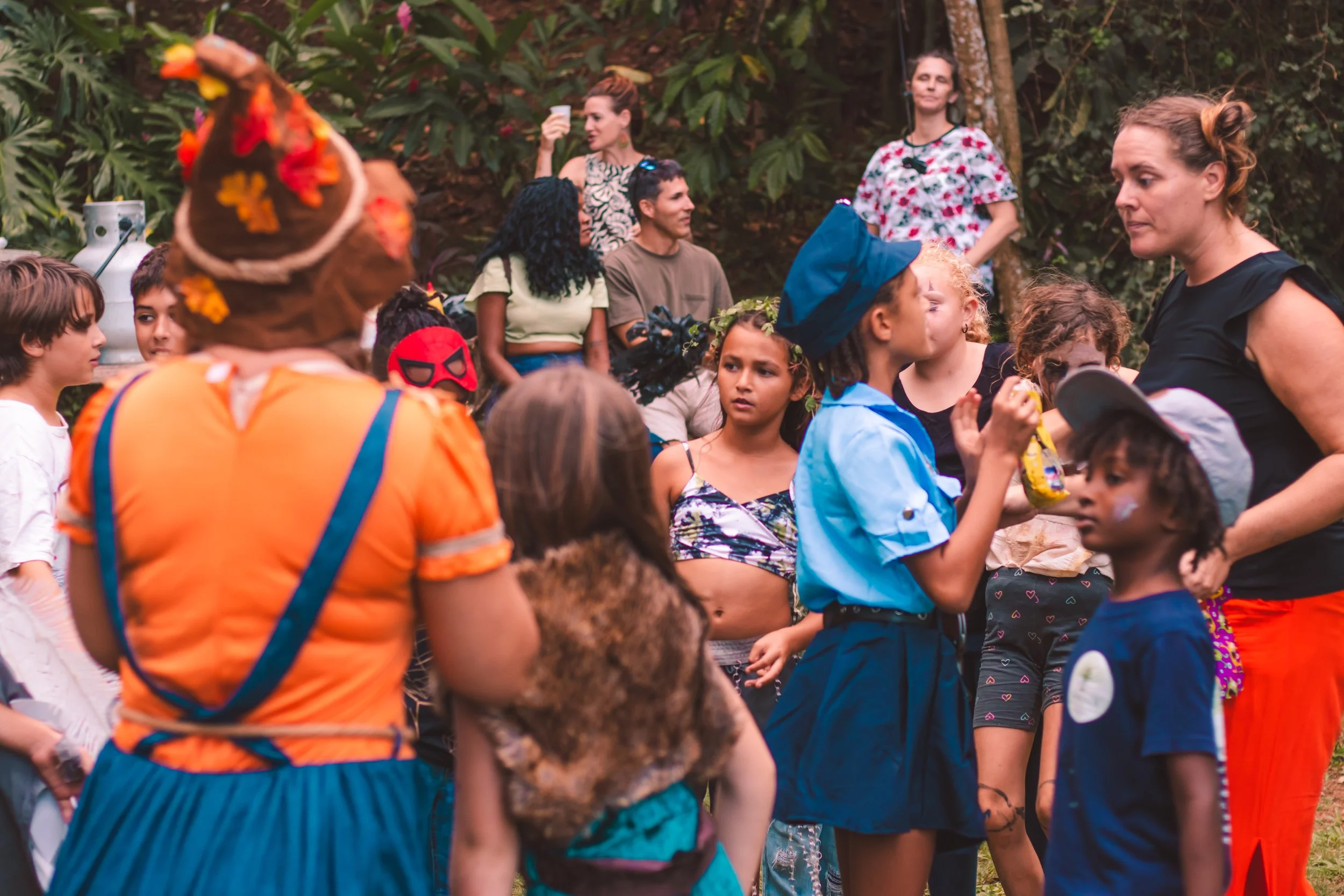 Children and adults gathered outdoors, some dressed in costumes, at a party or festival in a garden setting.