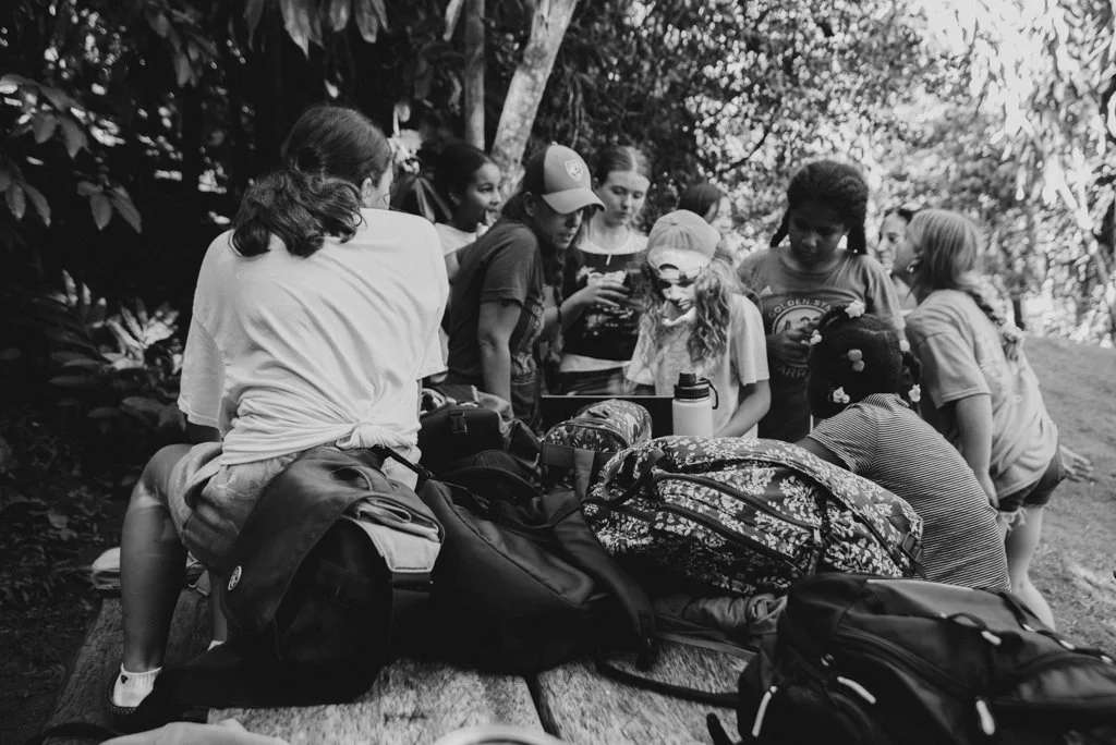 Group of children gathered outdoors around a table with backpacks and bags.