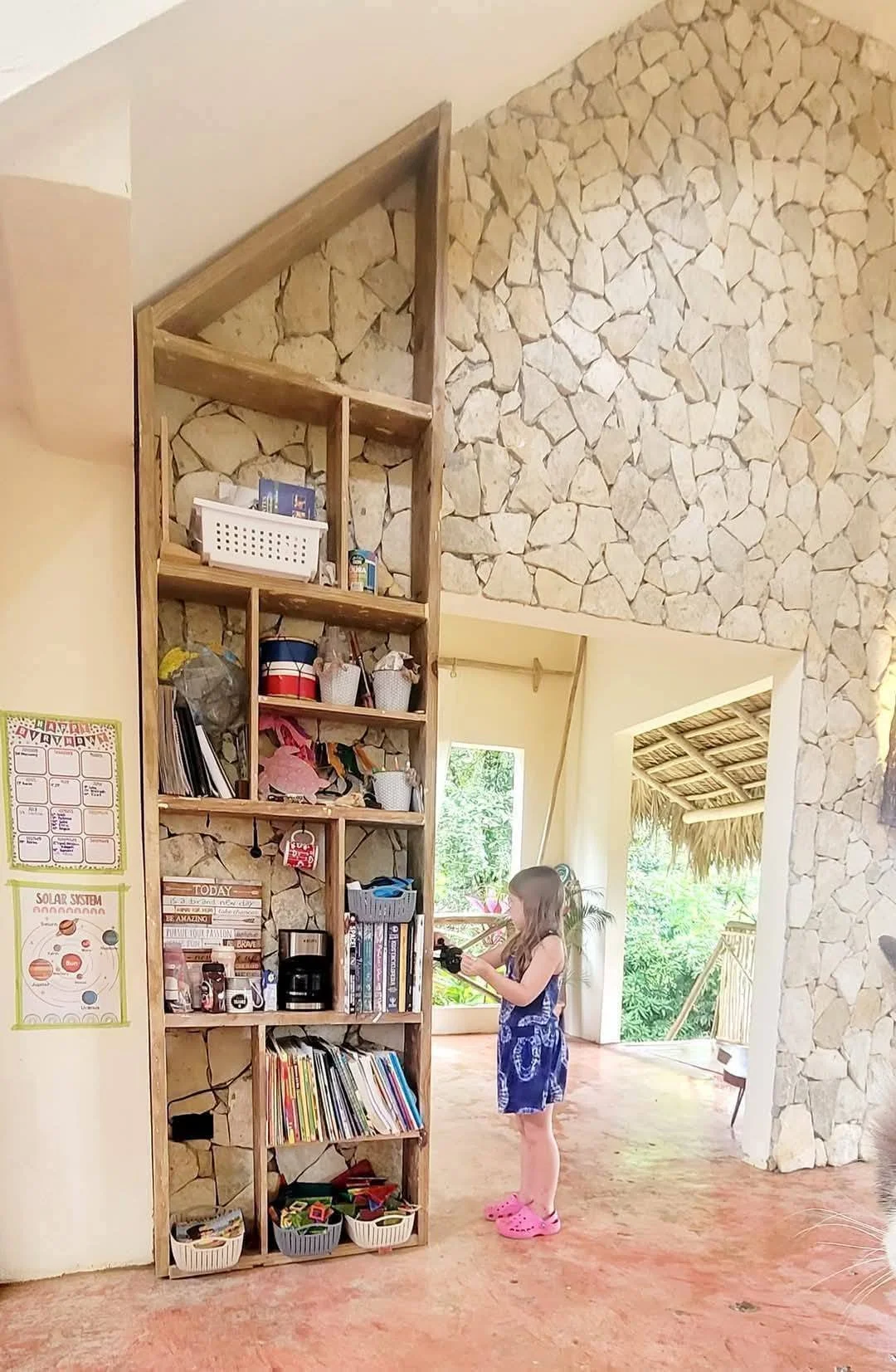A girl in a blue dress and pink Crocs, standing indoors near a tall wooden bookshelf against a stone wall, holding a camera.