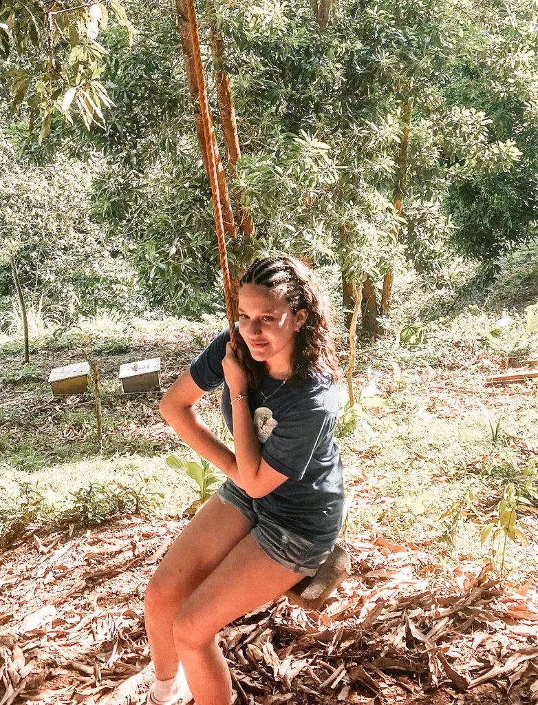 Young woman sitting on a swing made of a wooden seat and thick ropes, surrounded by lush green trees on a sunny day.