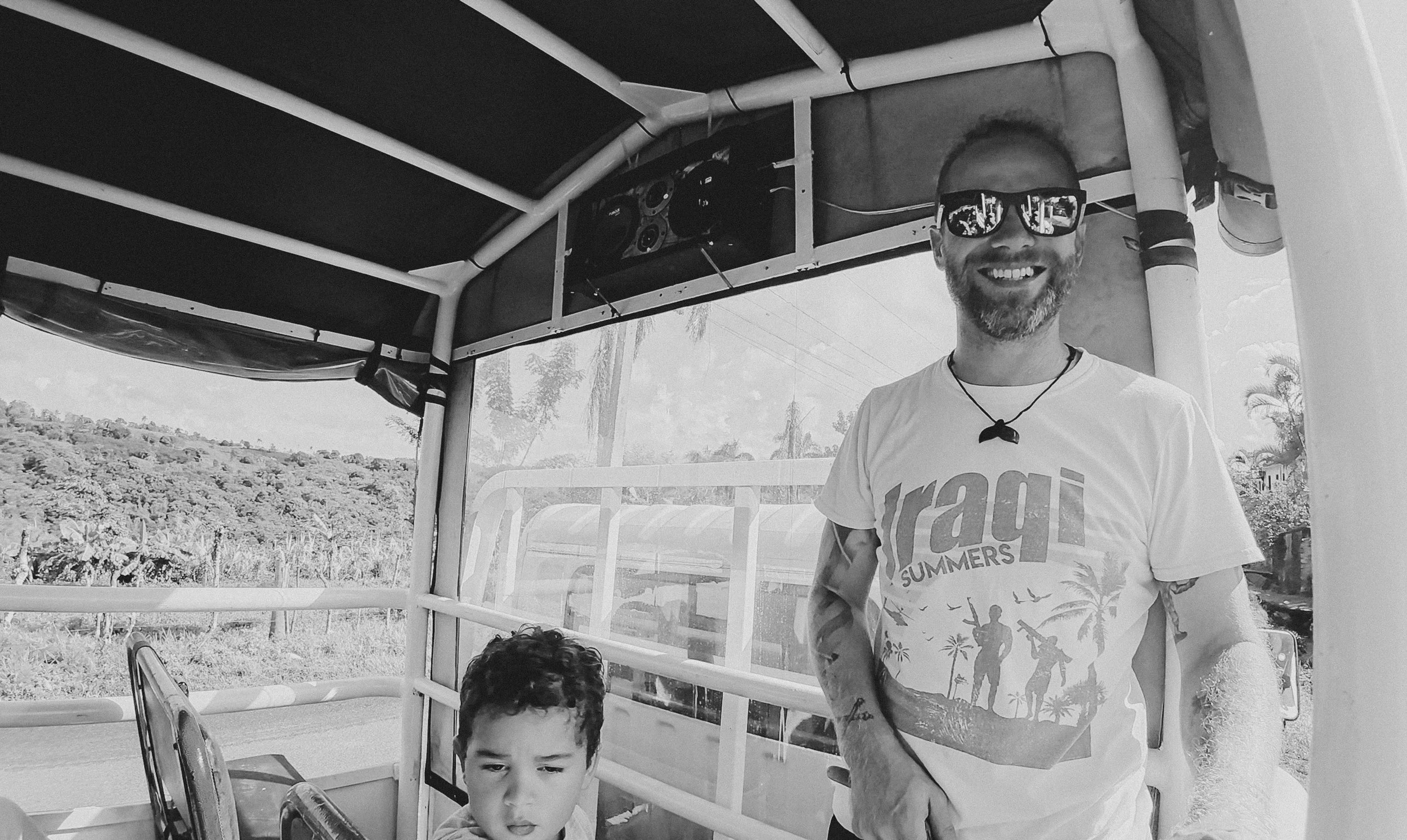 A man with a beard wearing sunglasses and a graphic summer T-shirt, smiling, standing next to a young boy with curly hair on a boat or tram, with a scenic landscape and trees in the background.