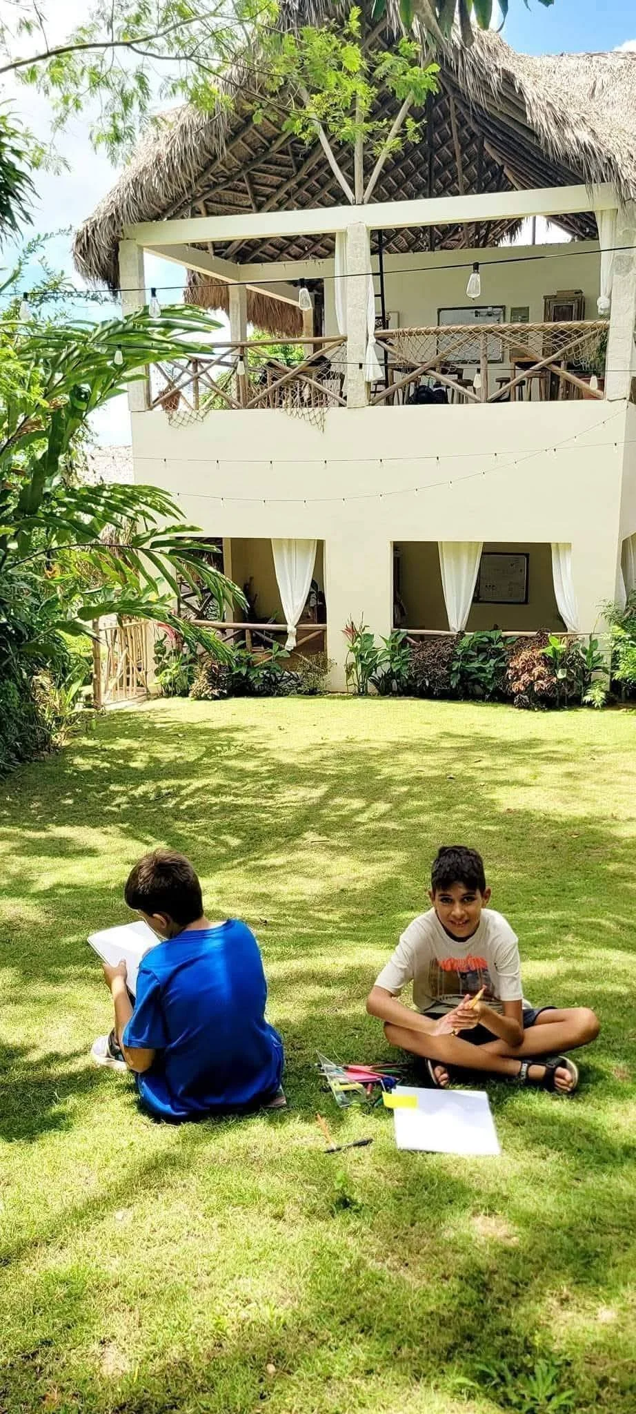Two boys sitting on a grassy lawn with art supplies, in front of a two-story house with an open balcony and tropical plants.