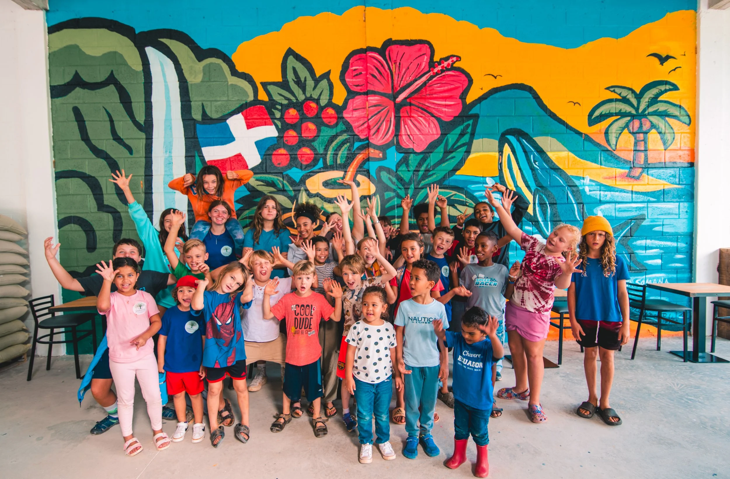 A group of children and a few adults smiling and posing for a photo in front of a colorful tropical mural with a mountain, waterfall, hibiscus flower, berries, and a palm tree.