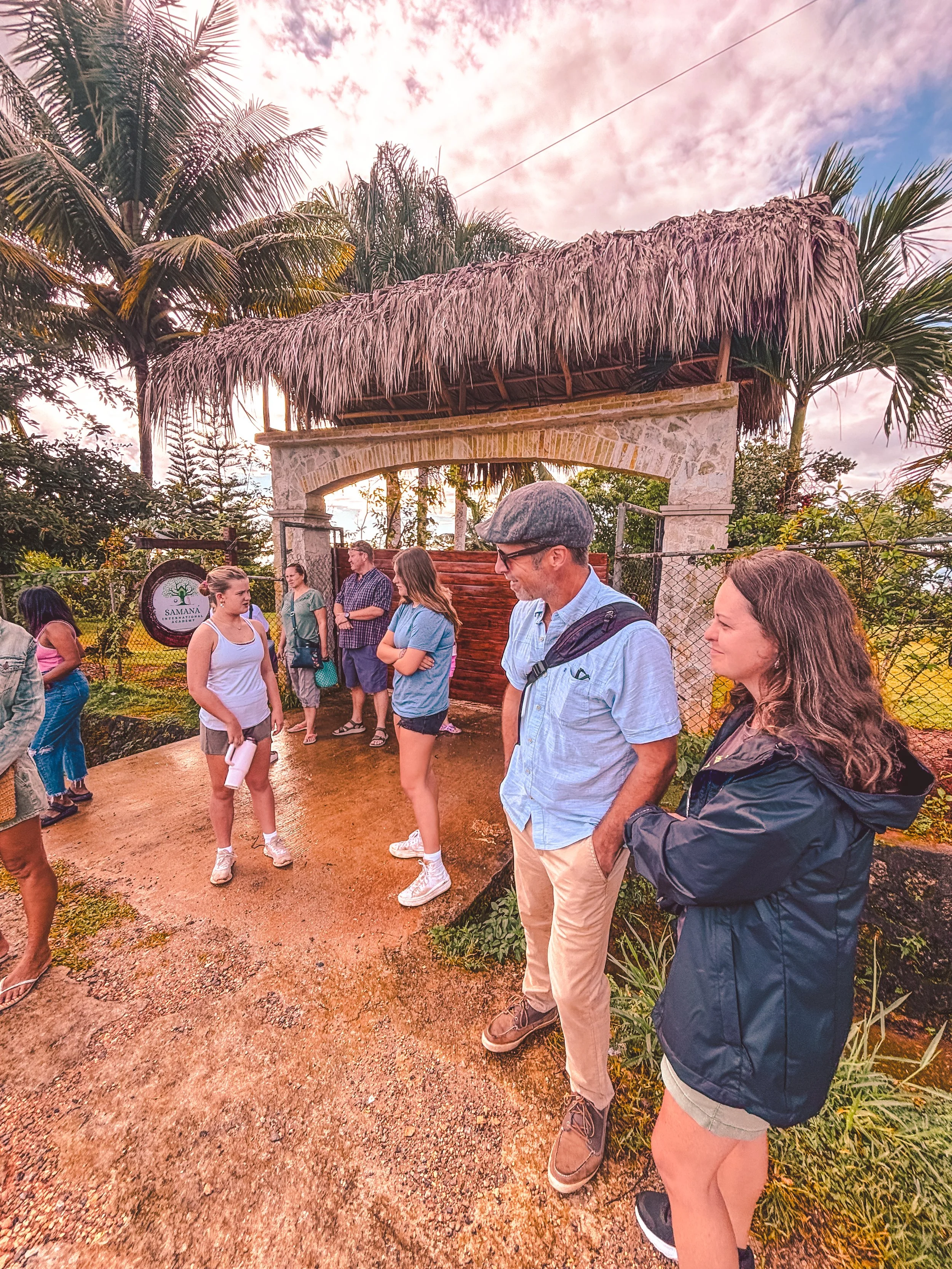 Group of people standing outdoors near tropical plants and a thatched roof entrance under a partly cloudy sky.