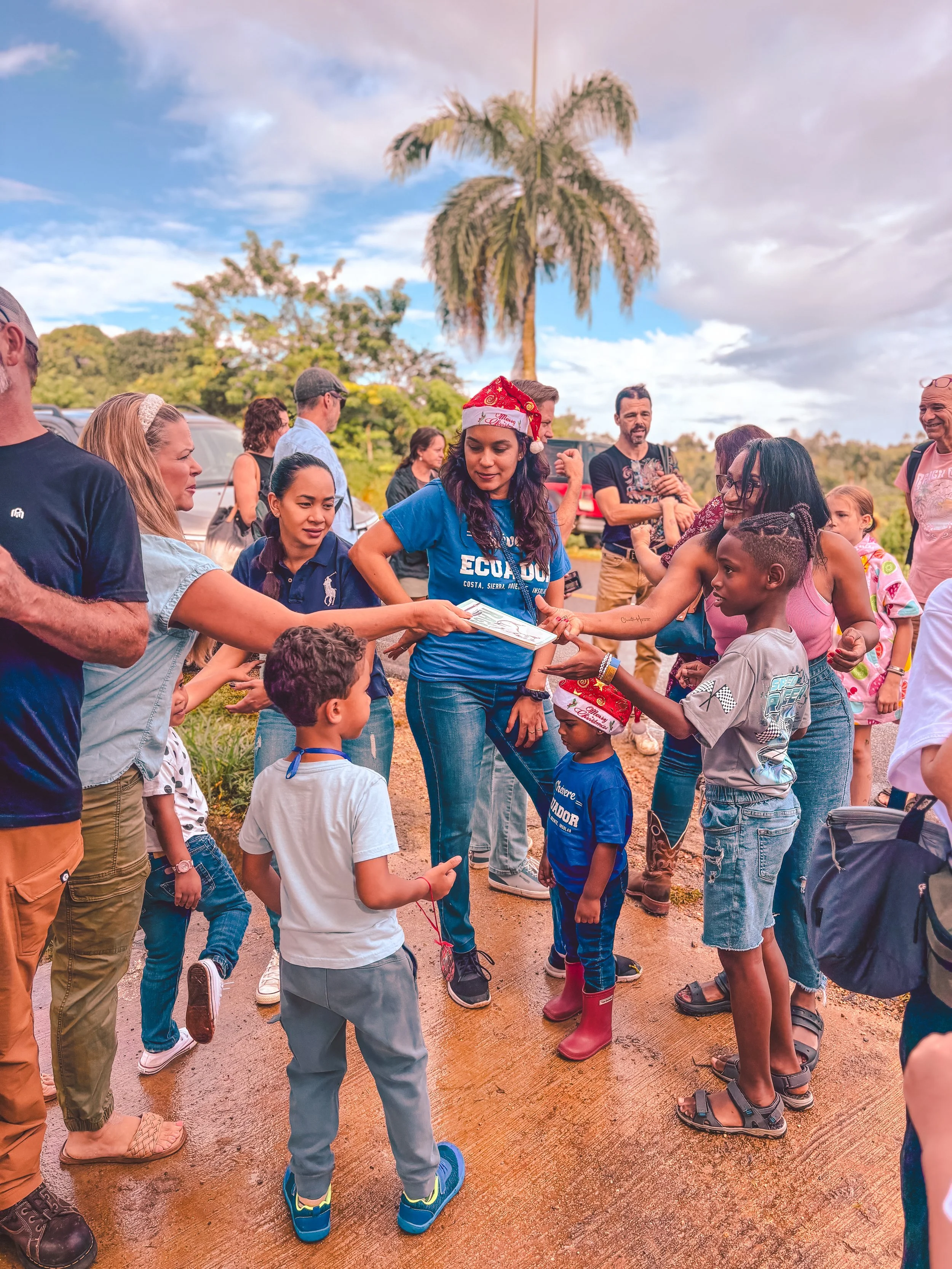 People gathered outdoors for a festive event, with children and adults exchanging gifts or awards, some wearing Christmas hats, under a cloudy sky with a tall palm tree in the background.