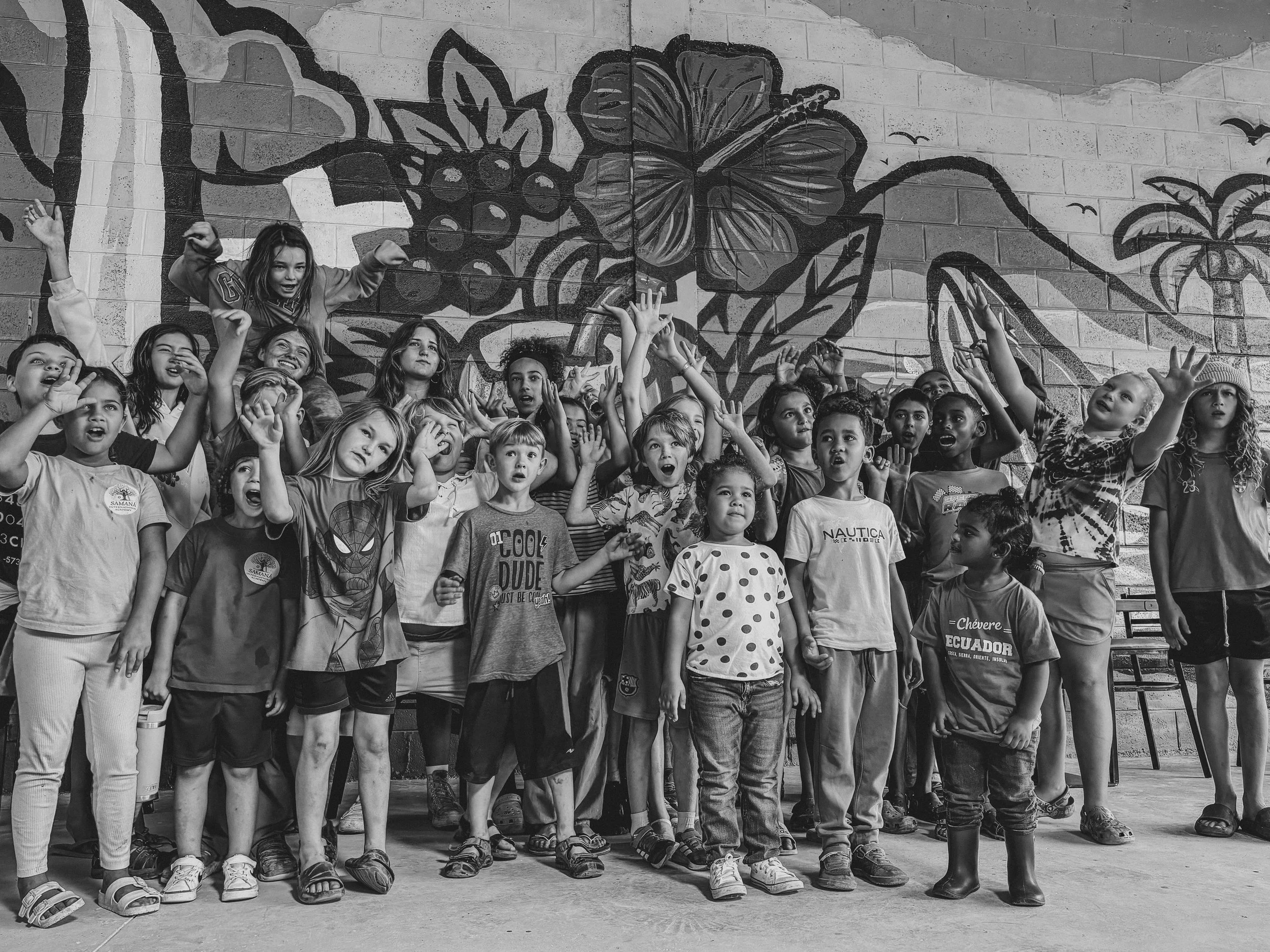 Group of children standing in front of a colorful mural, some raising their hands and making gestures, appearing to sing or perform.