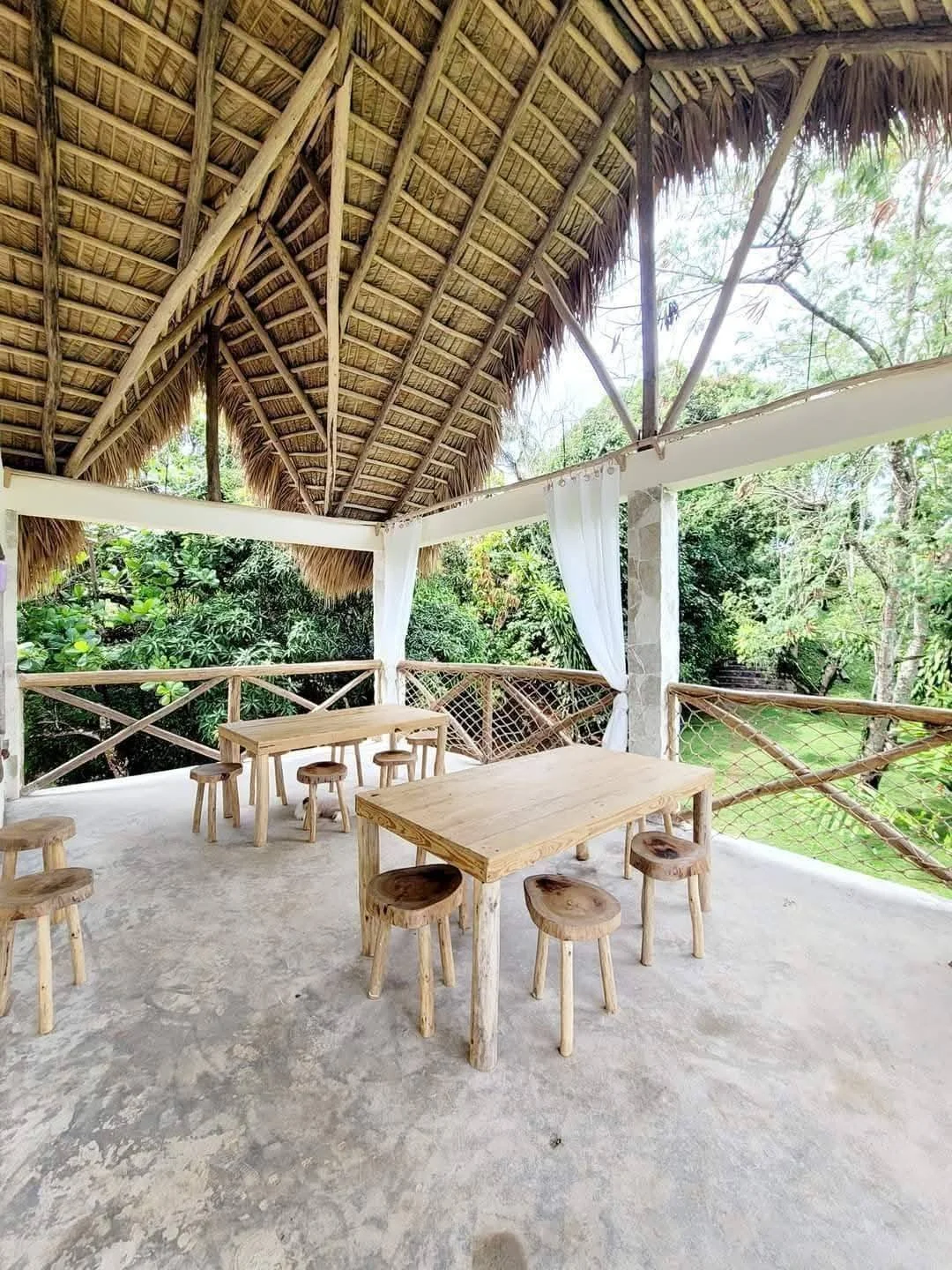Open-air tropical hut with thatched roof, wooden tables, small stools, and surrounding lush green foliage.