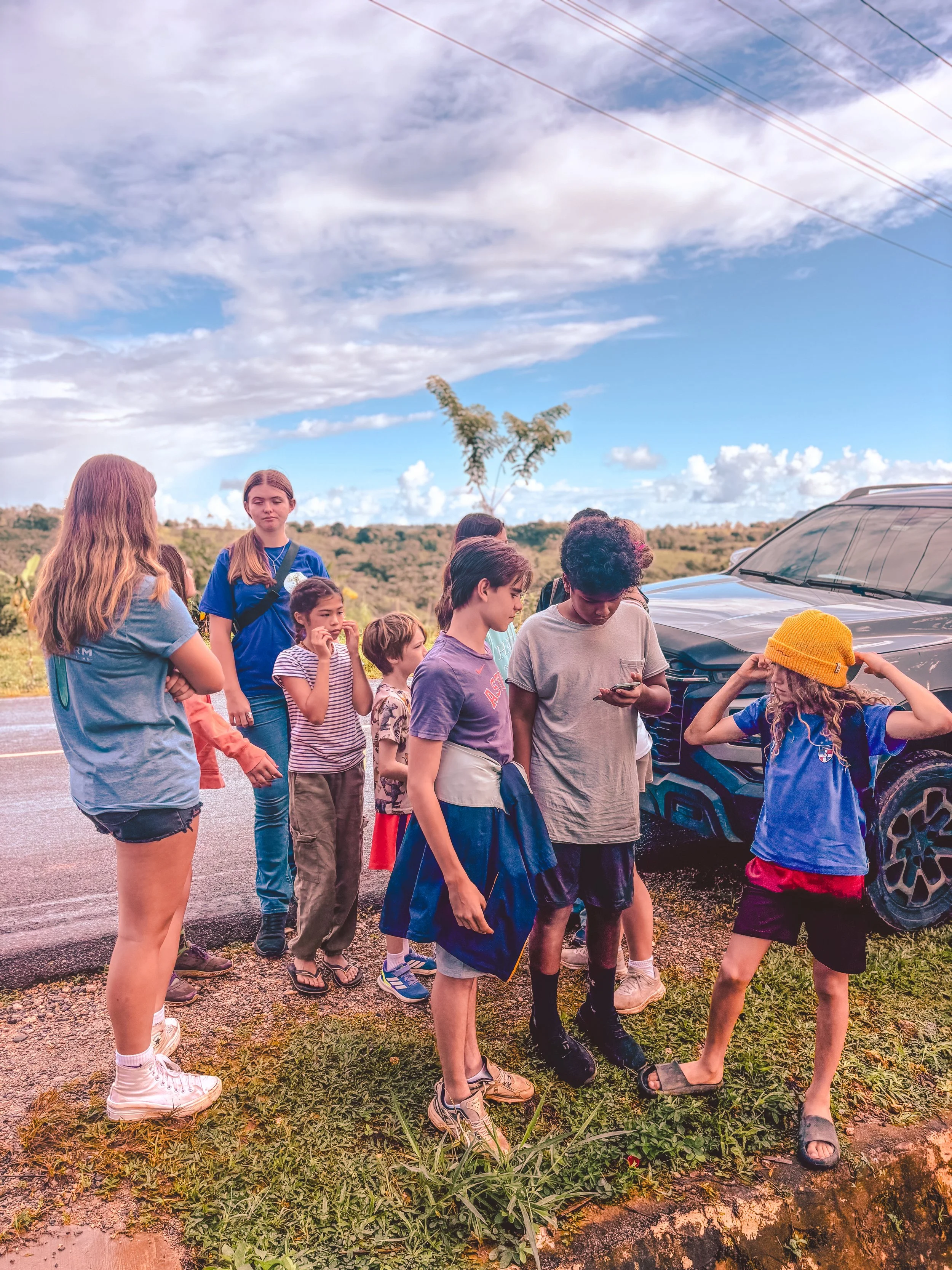Group of children and teenagers standing beside a black vehicle on a rural road, some looking at their phones, in a landscape with cloudy sky and trees in the background.
