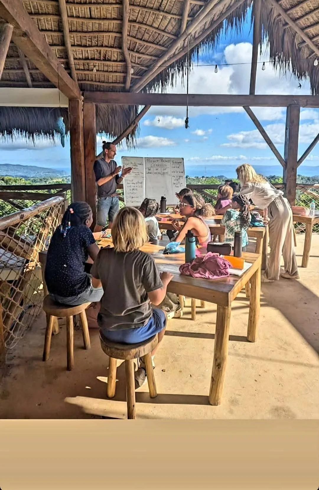 A group of children and adults attending an outdoor class in a thatched-roof open-air structure, with a teacher at a whiteboard and a scenic landscape of hills and blue sky in the background.