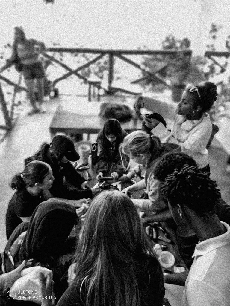 A group of women gathered around a table, looking at their phones or devices, in an outdoor setting with a wooden railing and trees in the background.