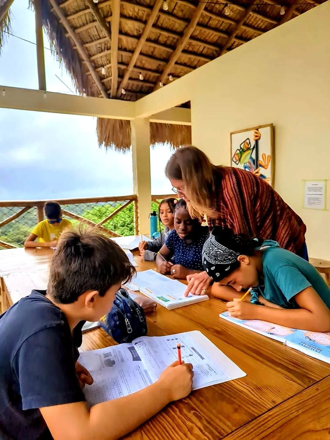 Children studying at a wooden table under a thatched roof, in an outdoor classroom setting, with a teacher assisting them.