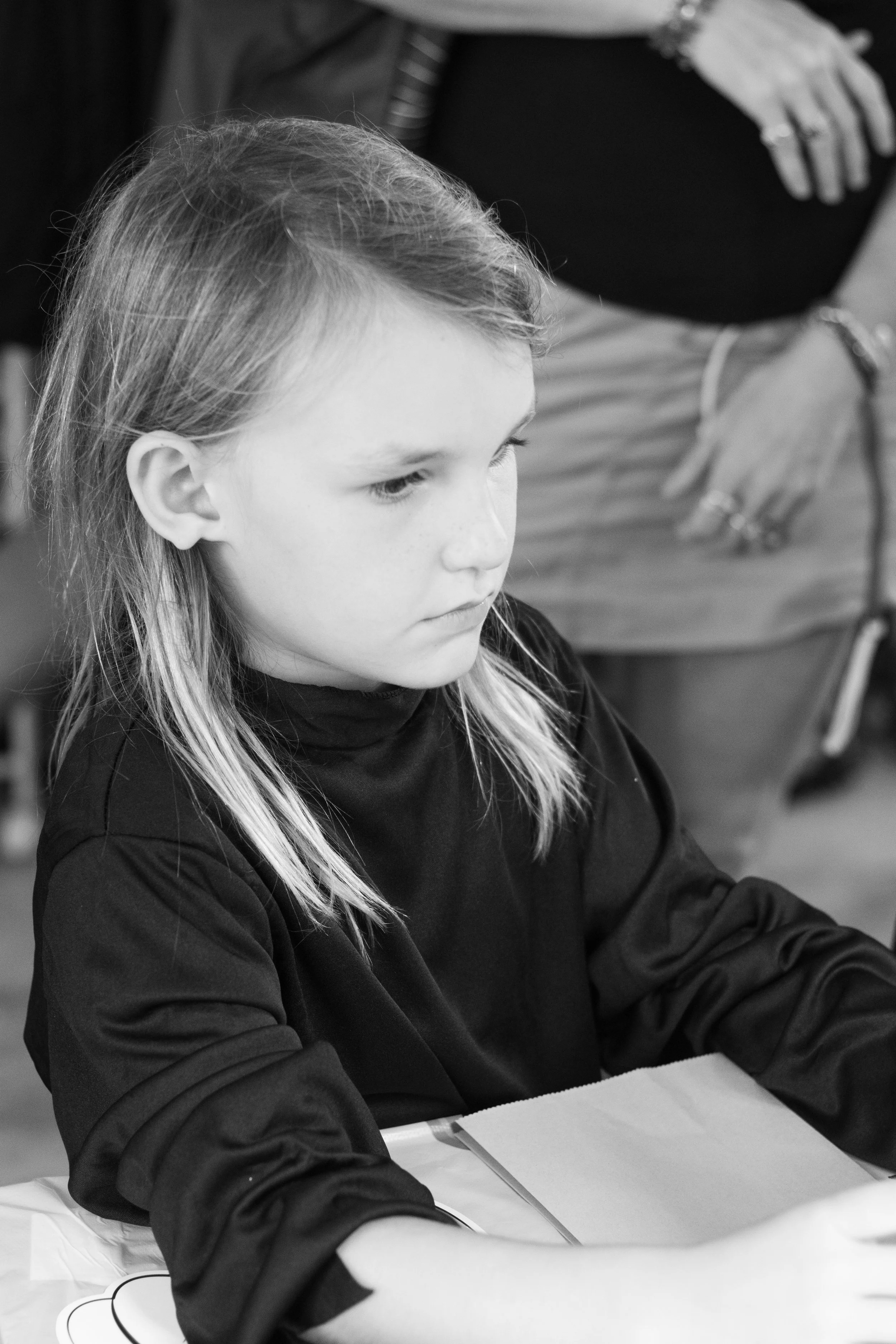 A young girl with long hair, wearing a dark shirt, seated at a table, appears focused, with an adult woman standing nearby.
