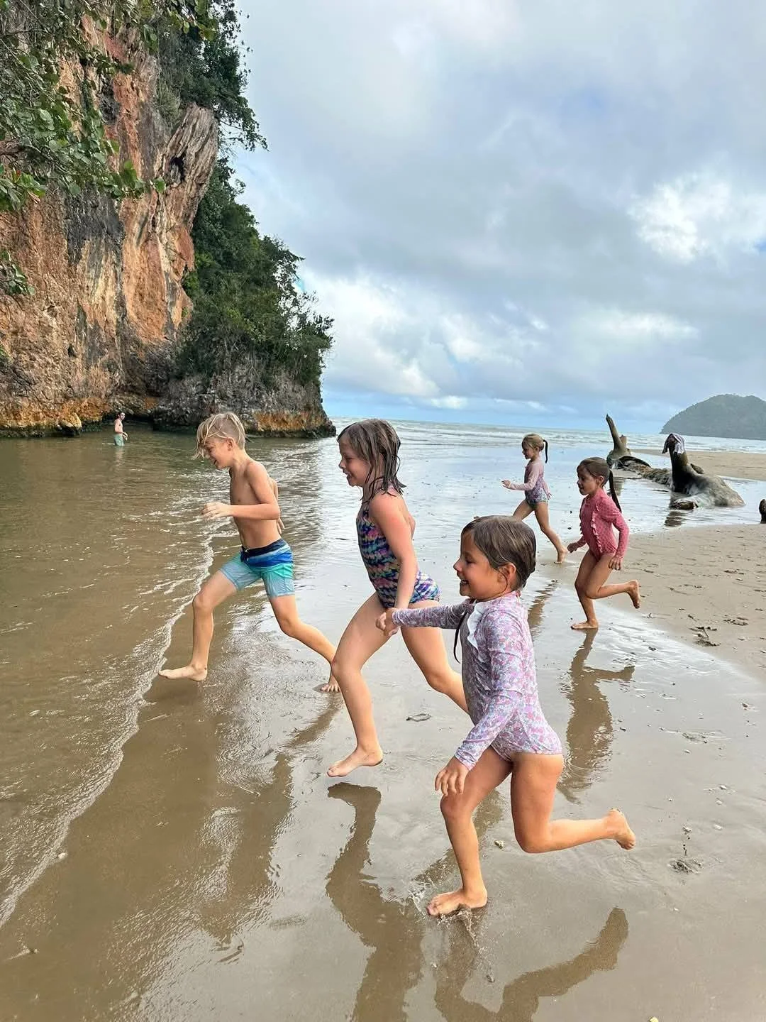 Children playing and running in shallow water on a beach with a rocky cliff and cloudy sky in the background.
