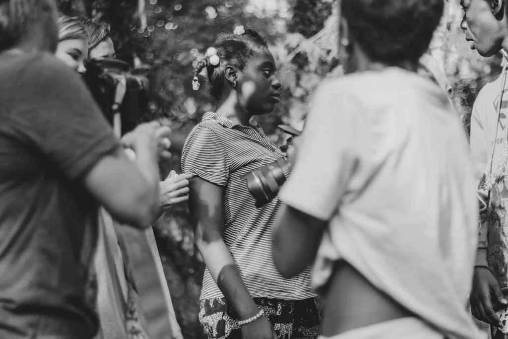 Group of people, mostly women, outdoors in a forested area, engaged in conversation, with one woman holding a camera.