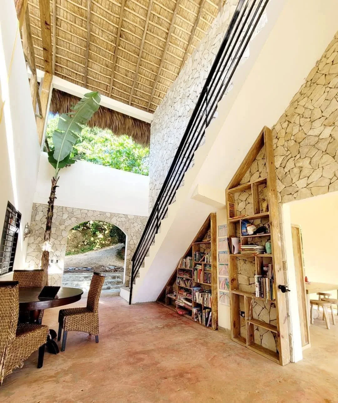 Interior view of a tropical house with high thatched roof, stone wall, wooden bookshelves, a round dining table with wicker chairs, a banana plant, and an arched doorway leading outside to greenery.