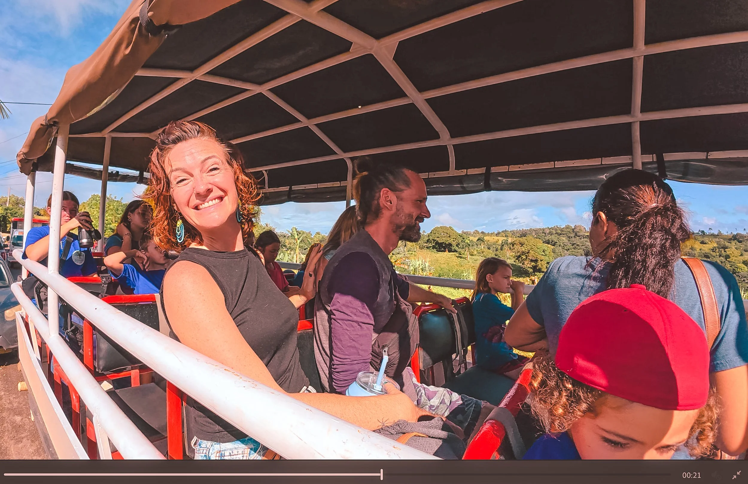 Smiling woman with curly red hair and turquoise earrings riding a bus with other passengers, including children, in a rural area with green trees and blue sky in the background.
