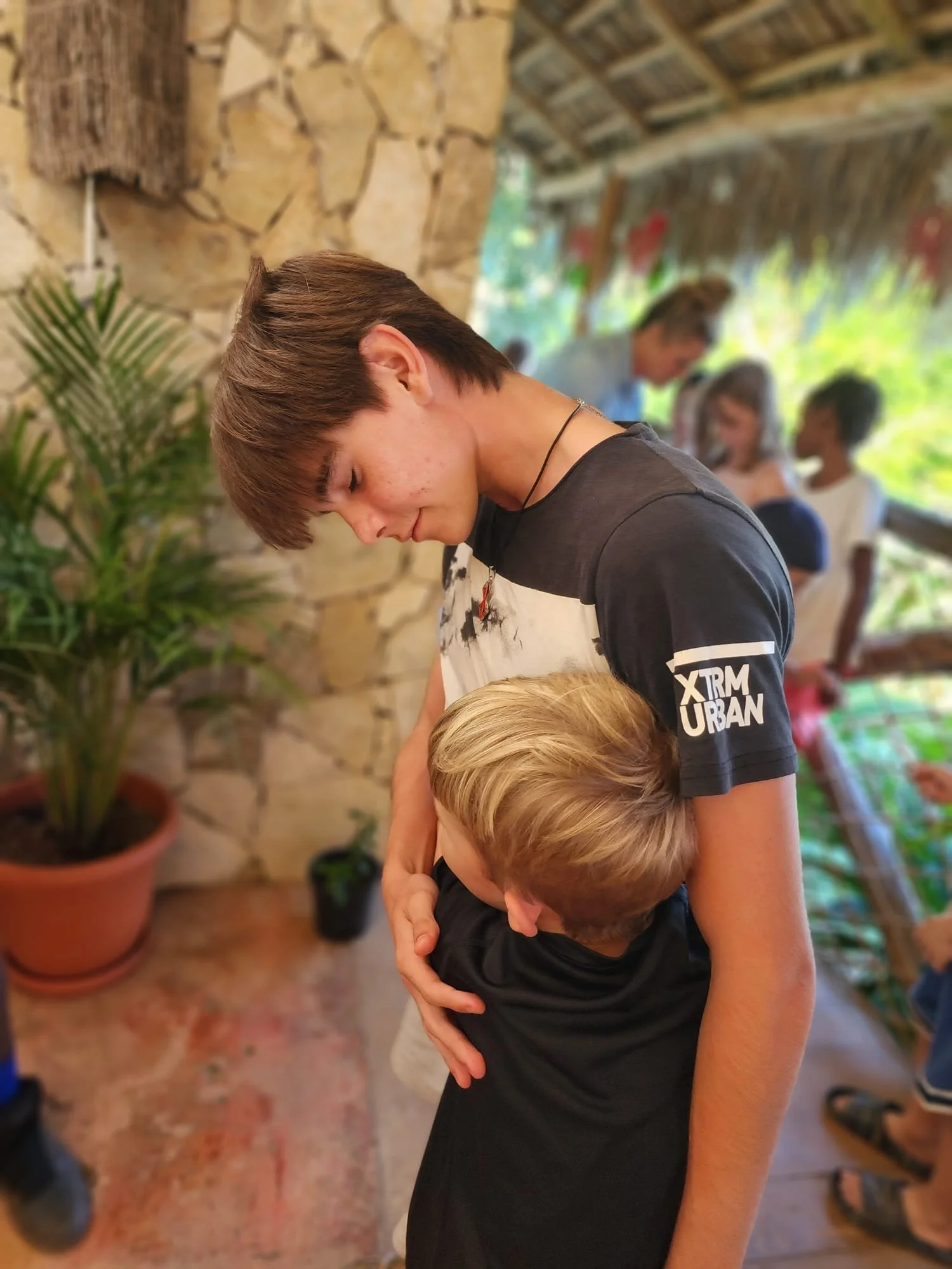 A teenage boy with short brown hair is gently hugging a young boy with blond hair. The scene takes place indoors with a stone wall and potted plants in the background. Several people are blurred in the background, including children.