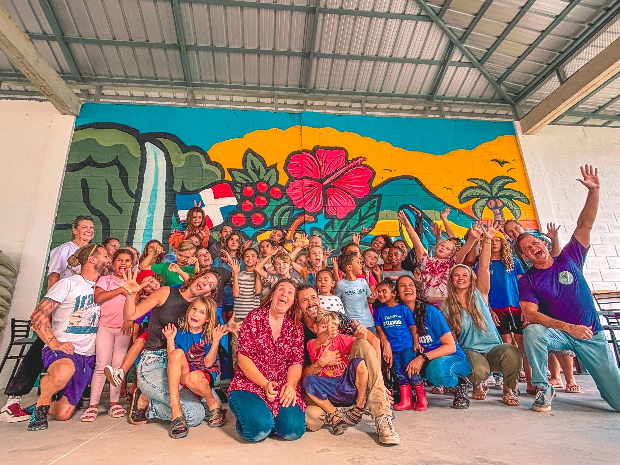 A group of smiling children and adults posing in front of a colorful mural featuring tropical scenery with a waterfall, hibiscus flower, and palm trees.