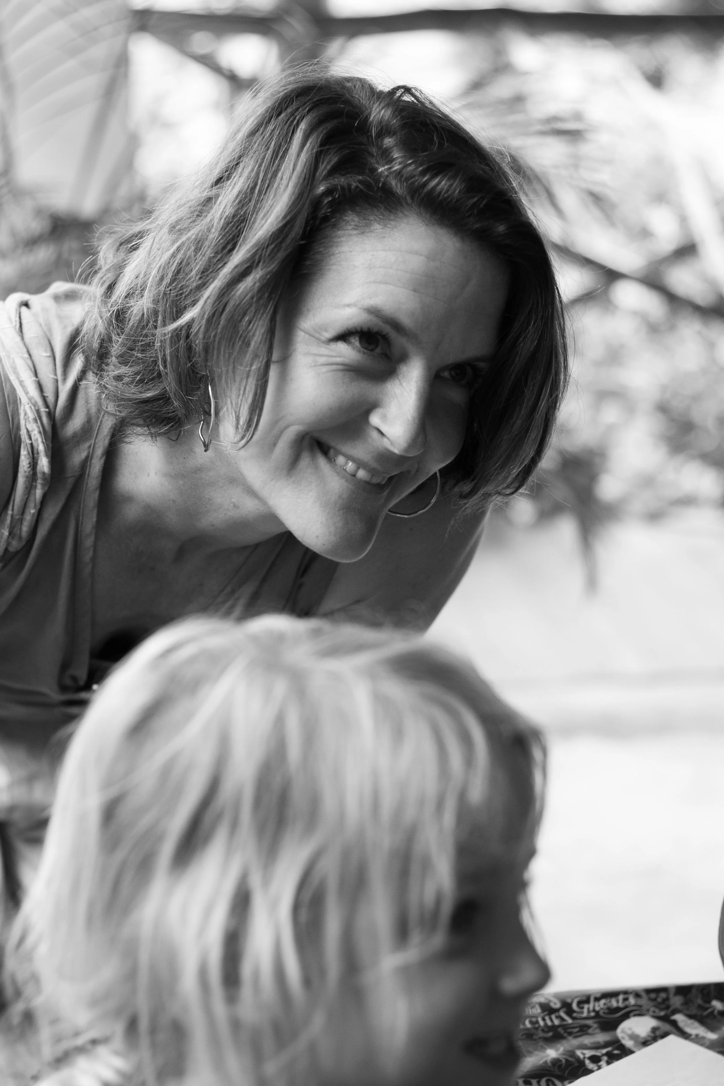 A woman with shoulder-length hair smiling and leaning toward a child with blonde hair, both outdoors.