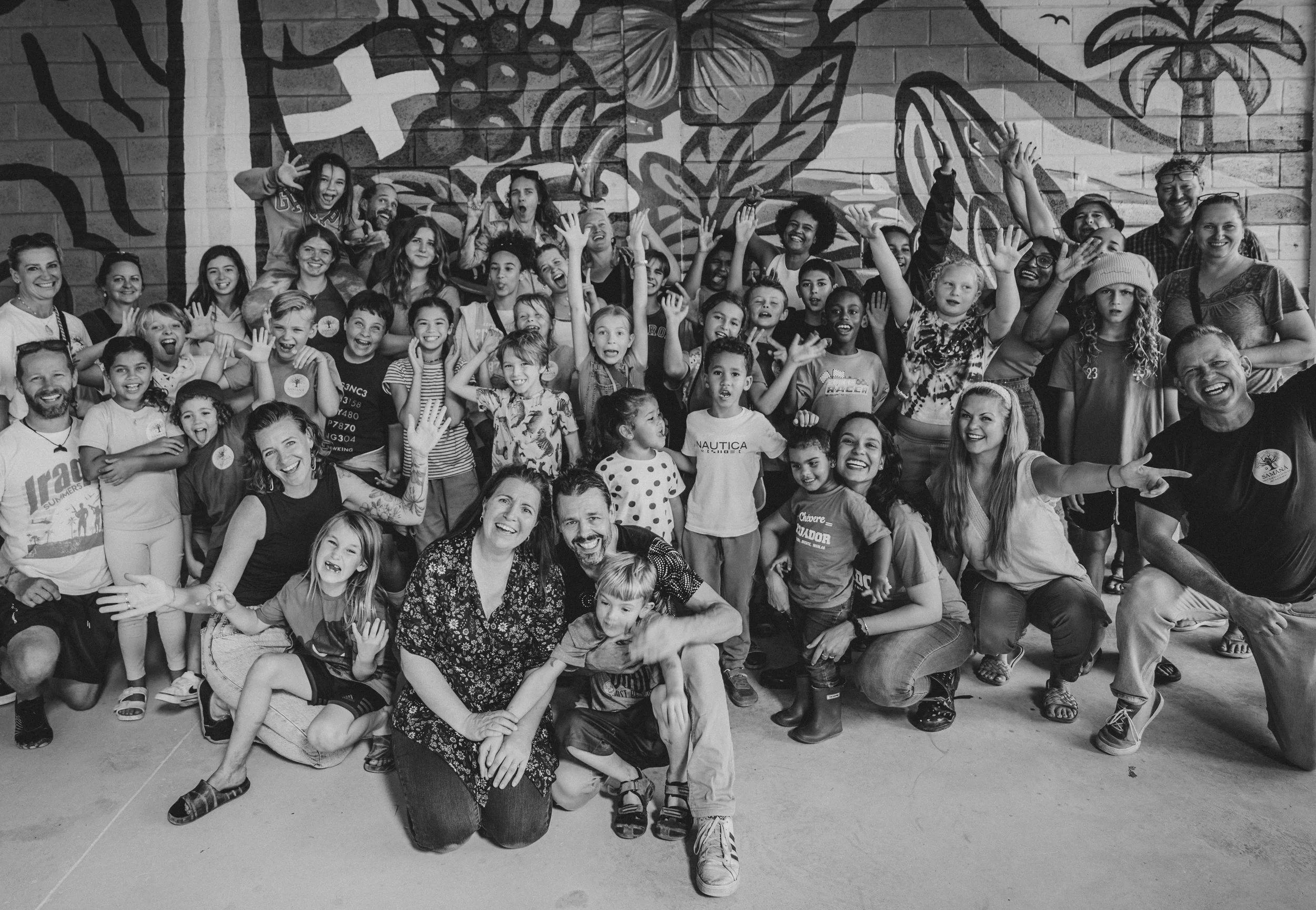 A large group of children and adults posing together in front of a mural wall, many are smiling, some are waving, and others are making playful gestures.