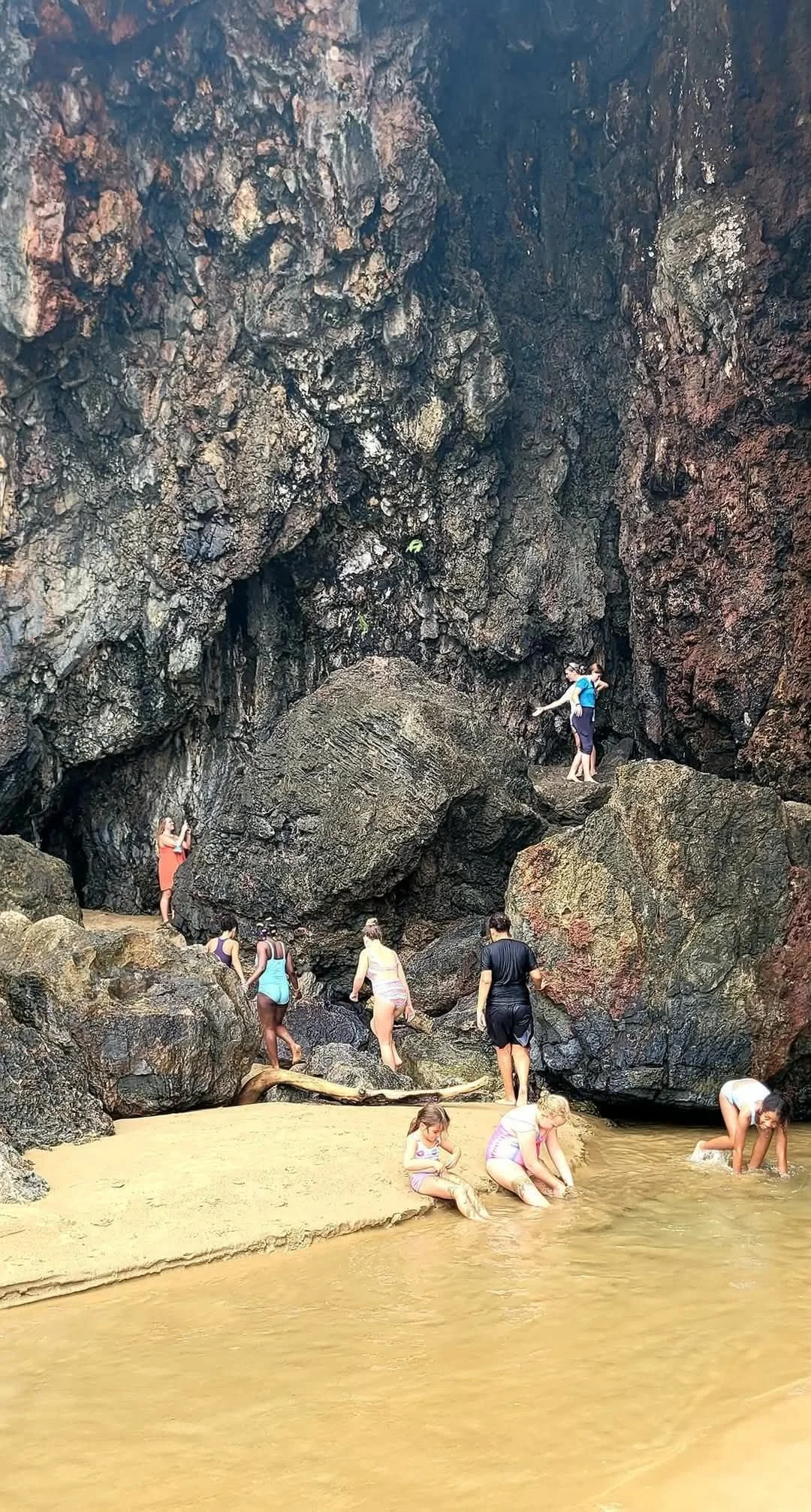 People exploring rocky terrain near water at a beach or cove with dark, rugged cliffs in the background.